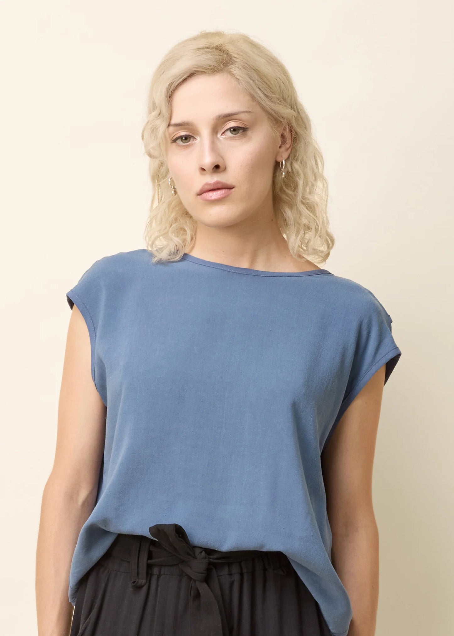 A woman wearing the Bayang Shirt by Cokluch in Ocean, featuring a round neck, short extended sleeves, and a cross-over bottom hem at the back. She is wearing it with black pants and is standing in front of a beige background.