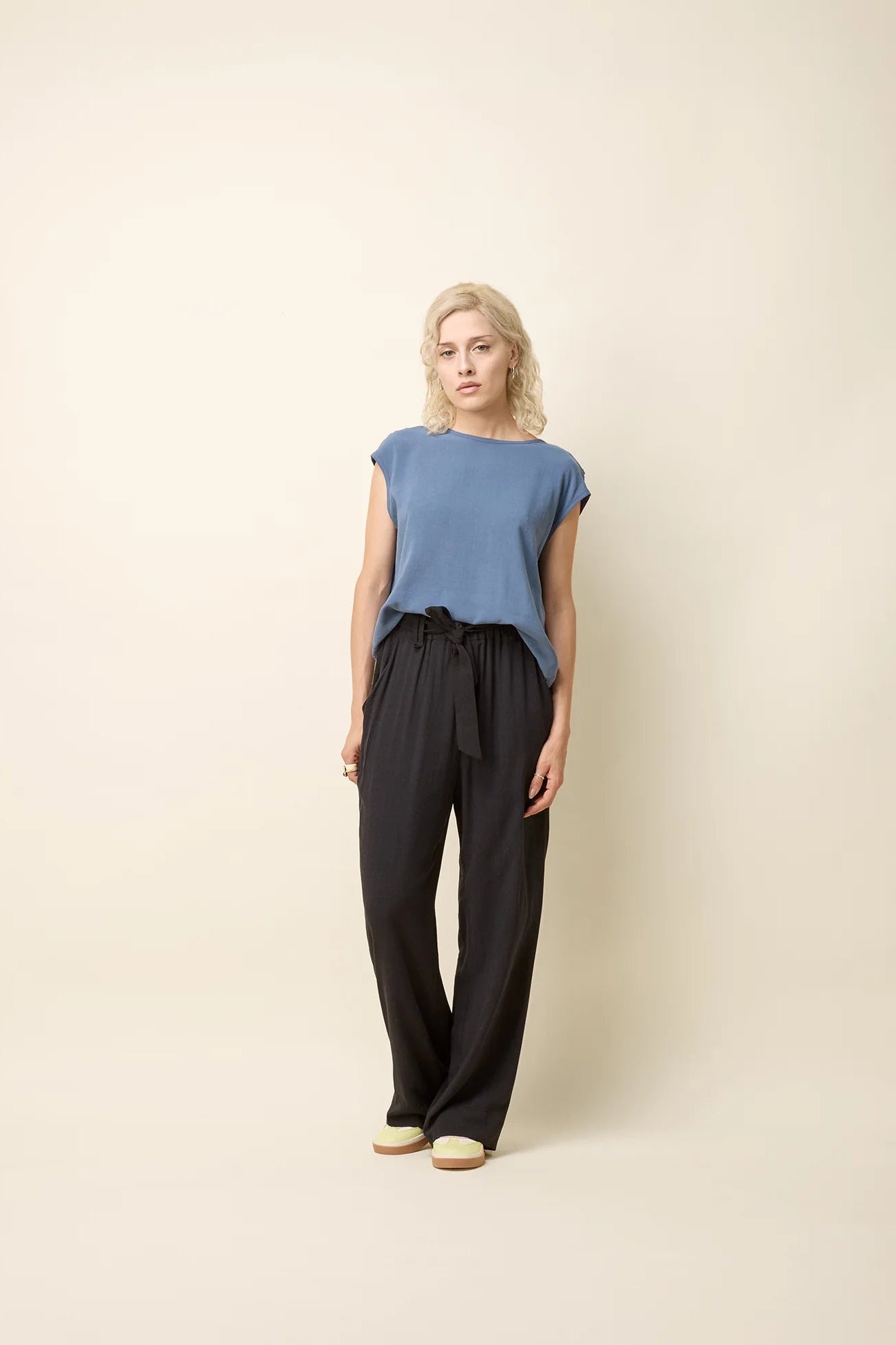 A woman wearing the Bayang Shirt by Cokluch in Ocean, featuring a round neck, short extended sleeves, and a cross-over bottom hem at the back. She is wearing it with black pants and is standing in front of a beige background.