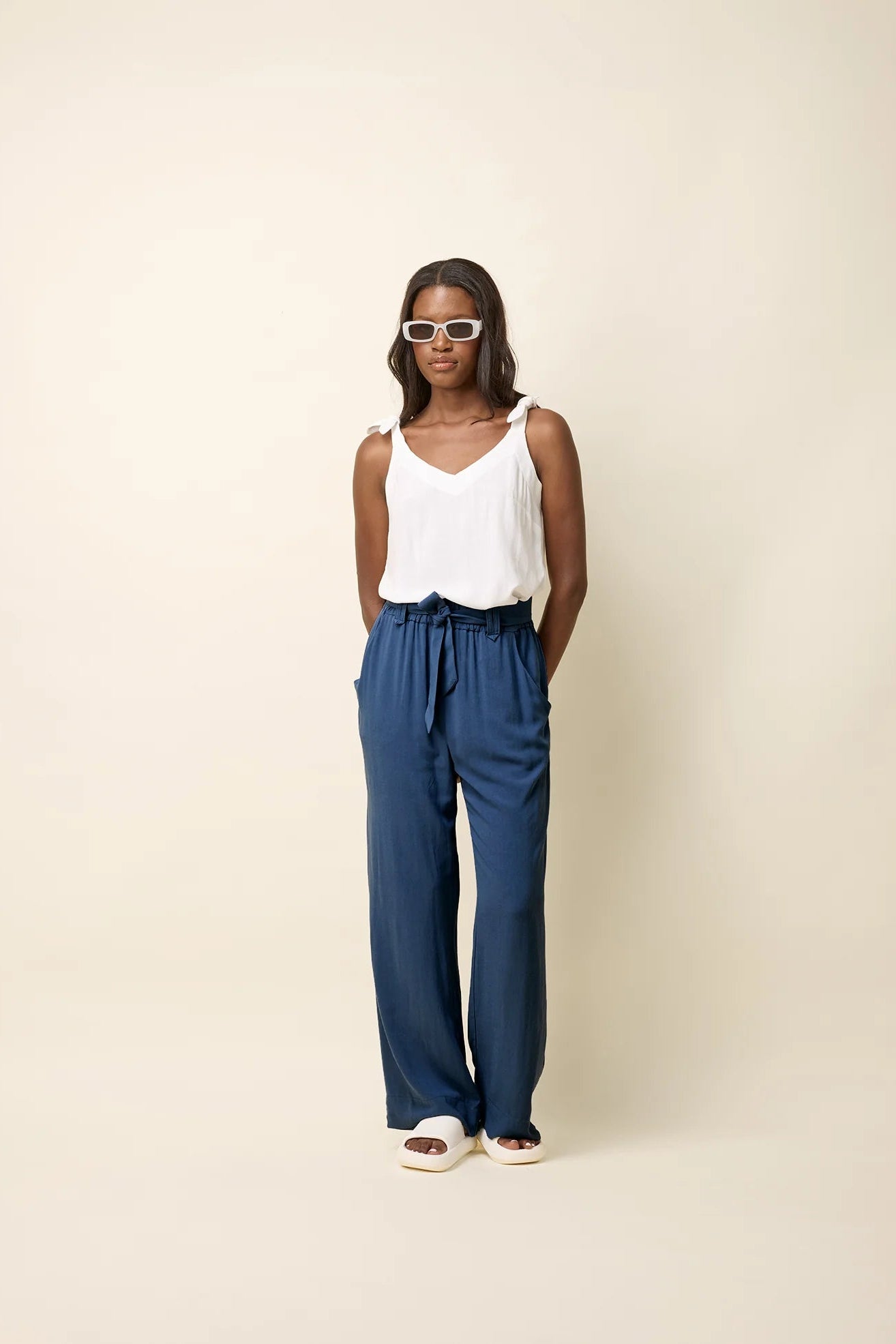 A woman wearing the Yael Pants by Cokluch in Ocean, slightly flared pants with an elastic waist, tie belt with pointed belt loops, and side pockets. She is wearing them with a white top and standing in front of a beige background.