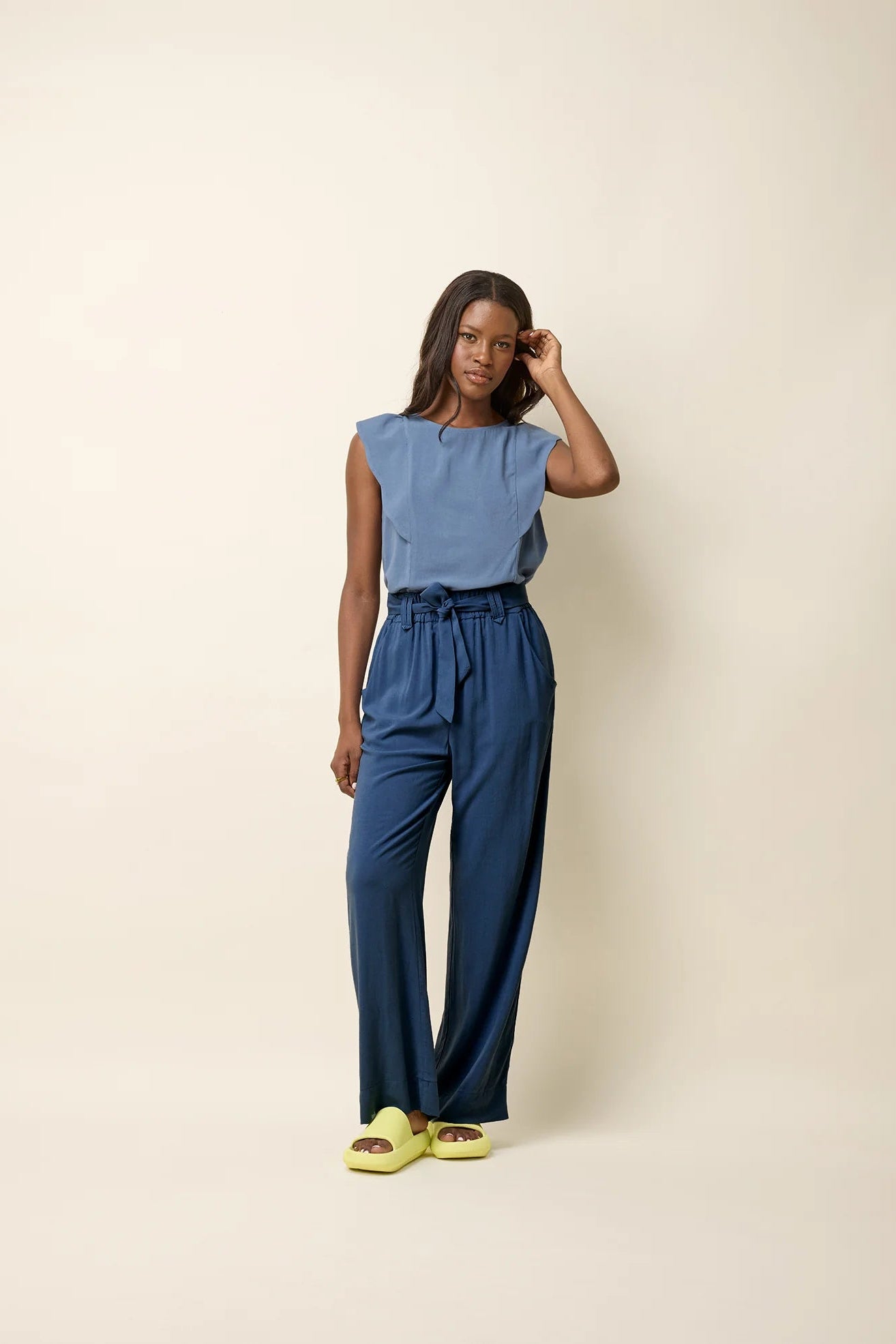 A woman wearing the Yael Pants by Cokluch in Ocean, slightly flared pants with an elastic waist, tie belt with pointed belt loops, and side pockets. She is wearing them with the Neema top in Ocean and standing in front of a beige background.