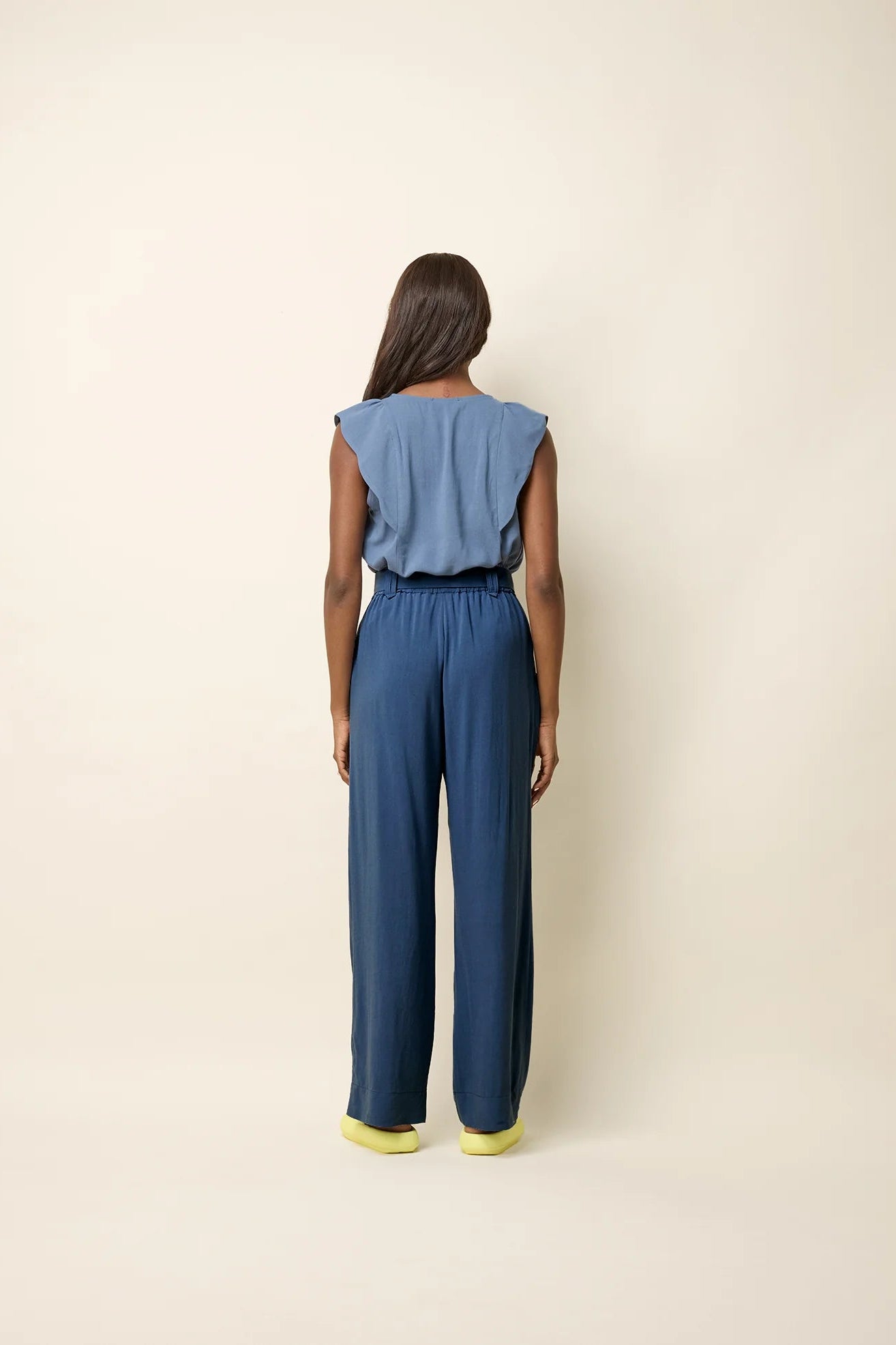 Back view of a woman wearing the Yael Pants by Cokluch in Ocean, slightly flared pants with an elastic waist, tie belt with pointed belt loops, and side pockets. She is wearing them with the Neema top in Ocean and standing in front of a beige background.