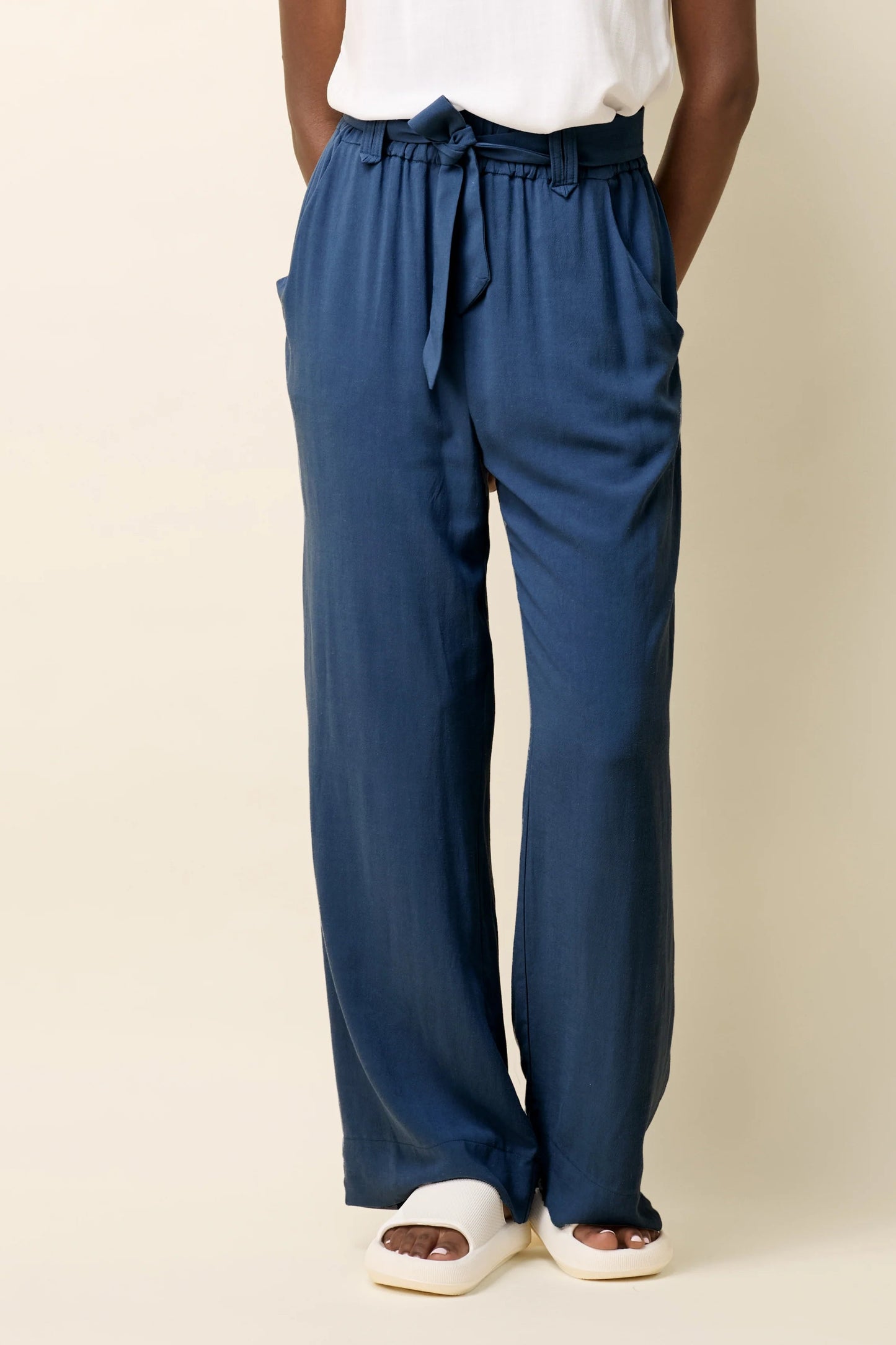 Waist down view of a woman wearing the Yael Pants by Cokluch in Ocean, slightly flared pants with an elastic waist, tie belt with pointed belt loops, and side pockets. She is wearing them with a white top and standing in front of a beige background.