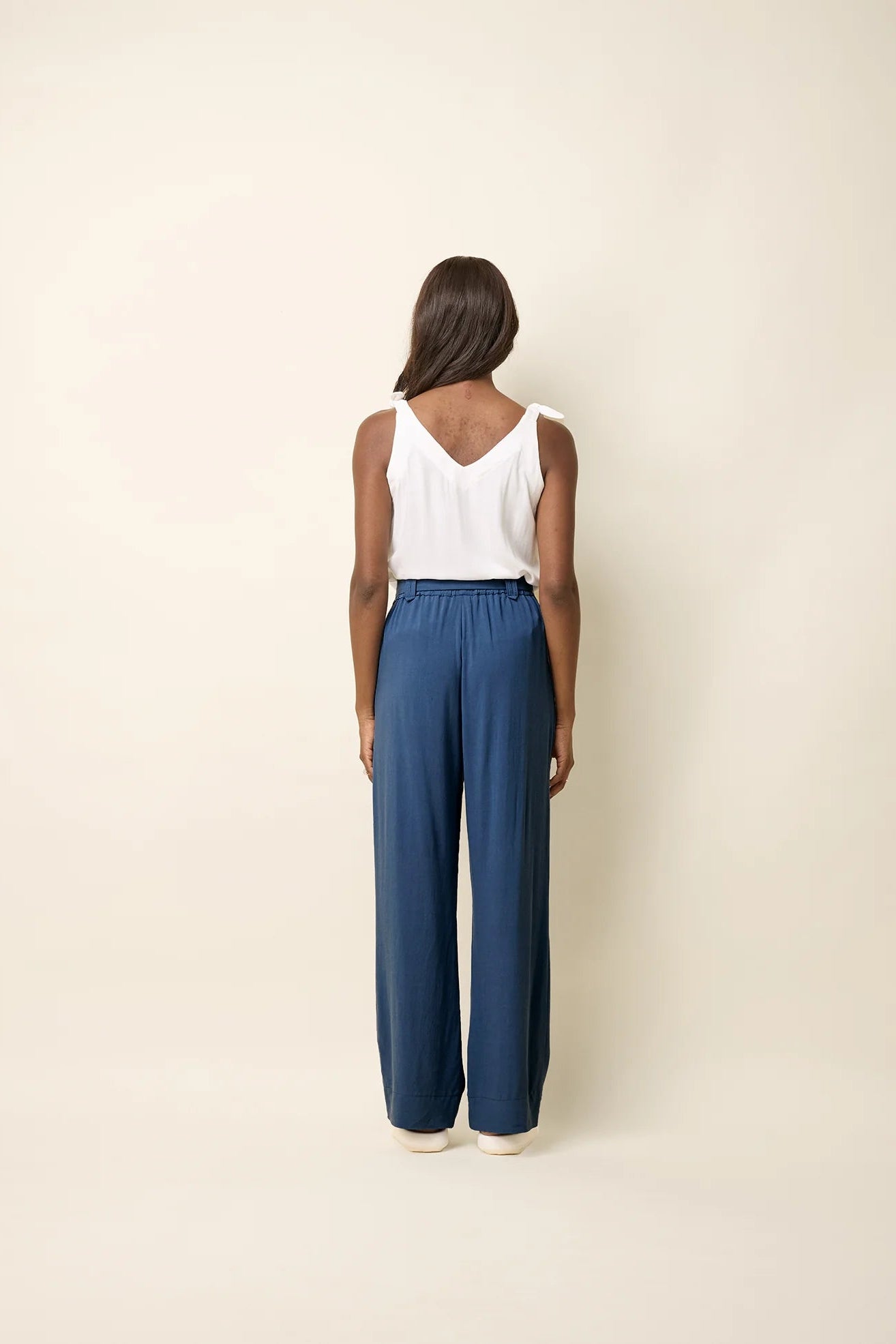 Back view of a woman wearing the Yael Pants by Cokluch in Ocean, slightly flared pants with an elastic waist, tie belt with pointed belt loops, and side pockets. She is wearing them with a white top and standing in front of a beige background.
