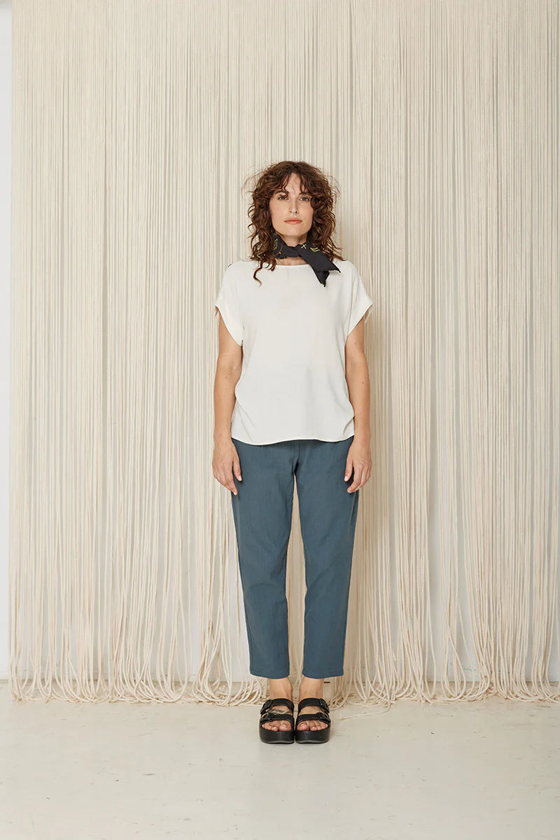 A woman wearing the Yasmina Pants from Cokluch in Steel, with a high elastic waist with a ring belt, and tapered, ankle-length legs. She is wearing them with a white top and standing in front of a beige curtain.