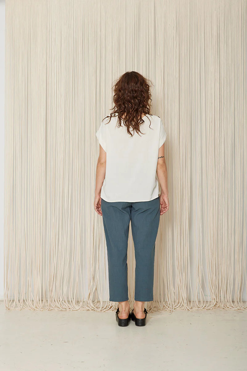Back view of a woman wearing the Yasmina Pants from Cokluch in Steel, with a high elastic waist with a ring belt, and tapered, ankle-length legs. She is wearing them with a white top and standing in front of a beige curtain.
