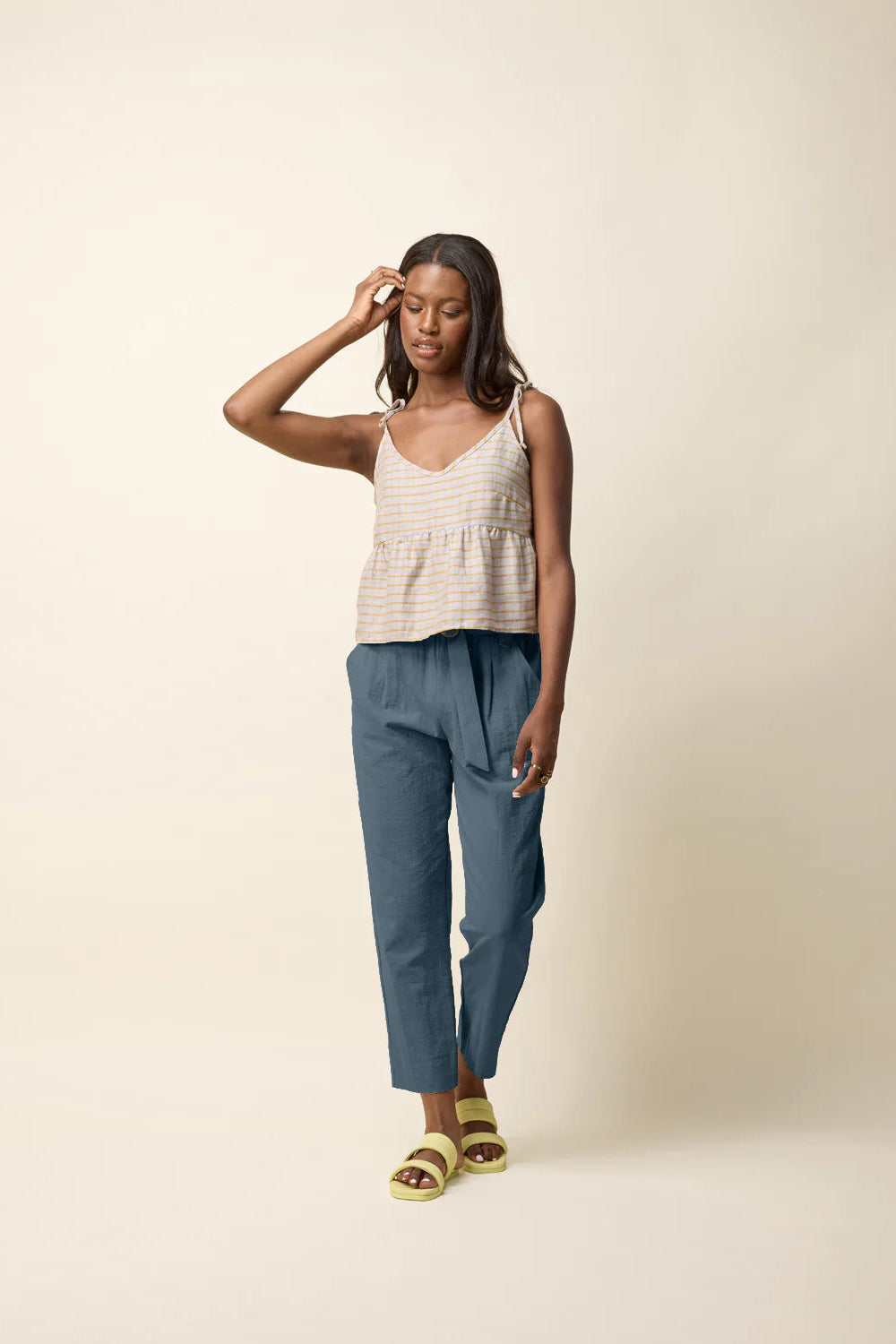 A woman wearing the Yasmina Pants from Cokluch in Steel, with a high elastic waist with a ring belt, and tapered, ankle-length legs. She is wearing them with a beige tank and standing in front of a beige background.