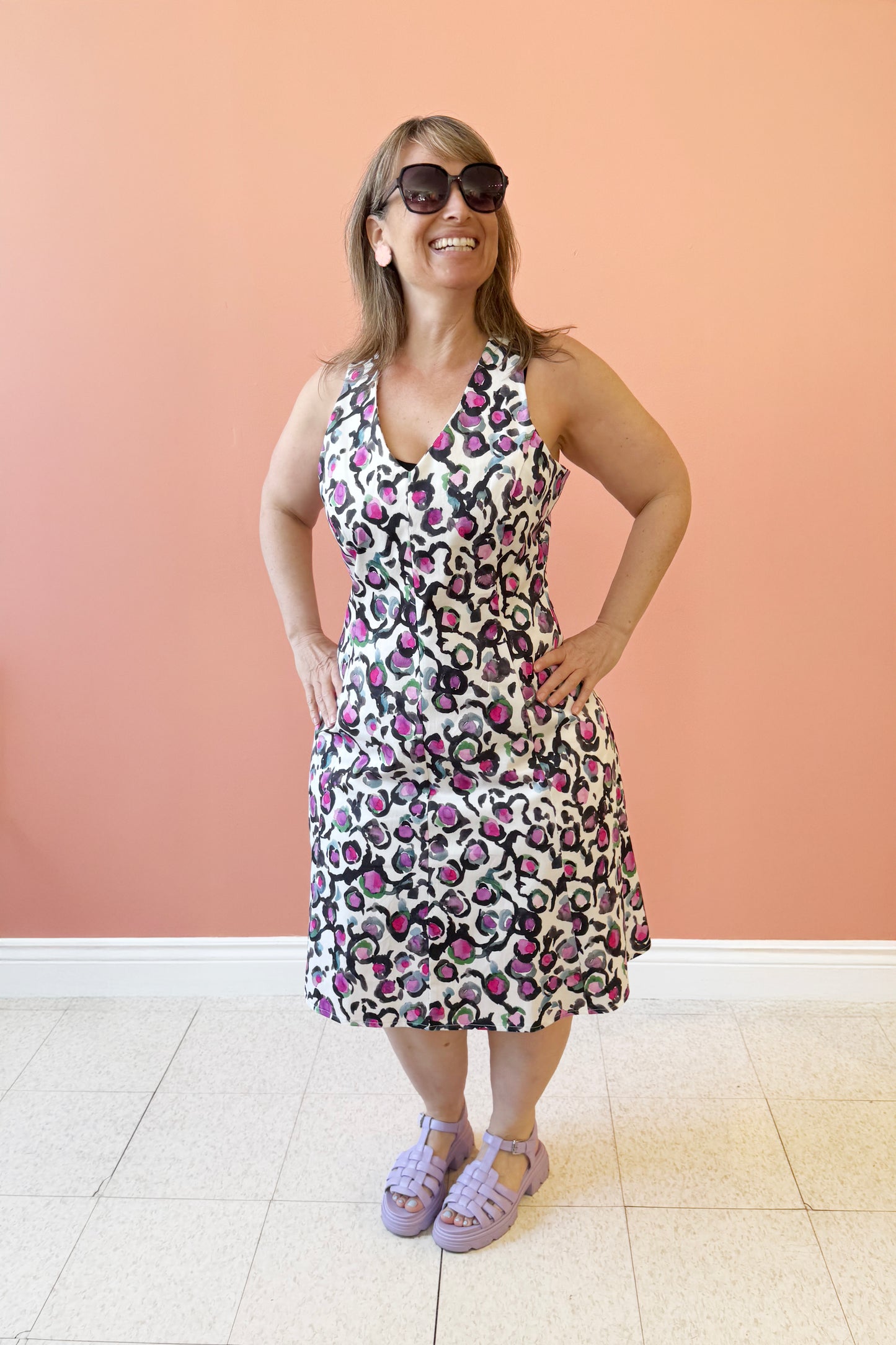 Bridget is wearing the Oceana Sundress by Compli K, an Abstract Floral sundress with a V-neck, wide straps, an A-line shape and a zip up the back. It falls to the knee. She's standing in front of a coral wall.