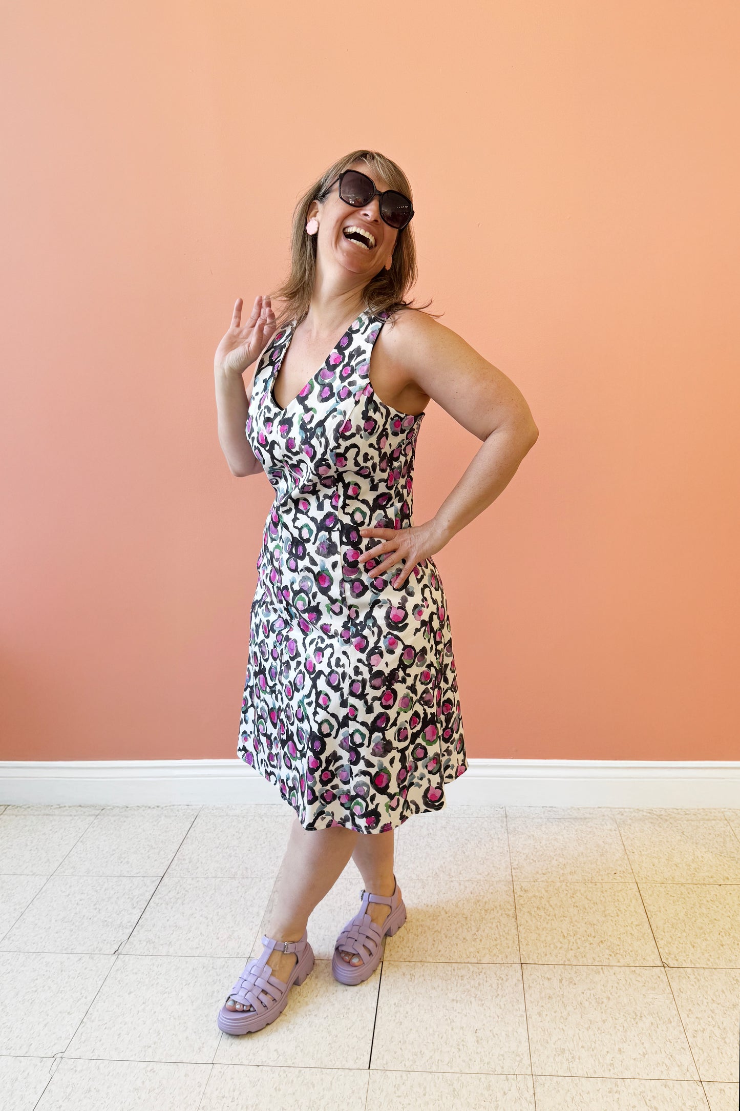 Bridget is wearing the Oceana Sundress by Compli K, an Abstract Floral sundress with a V-neck, wide straps, an A-line shape and a zip up the back. It falls to the knee. She's standing in front of a coral wall.