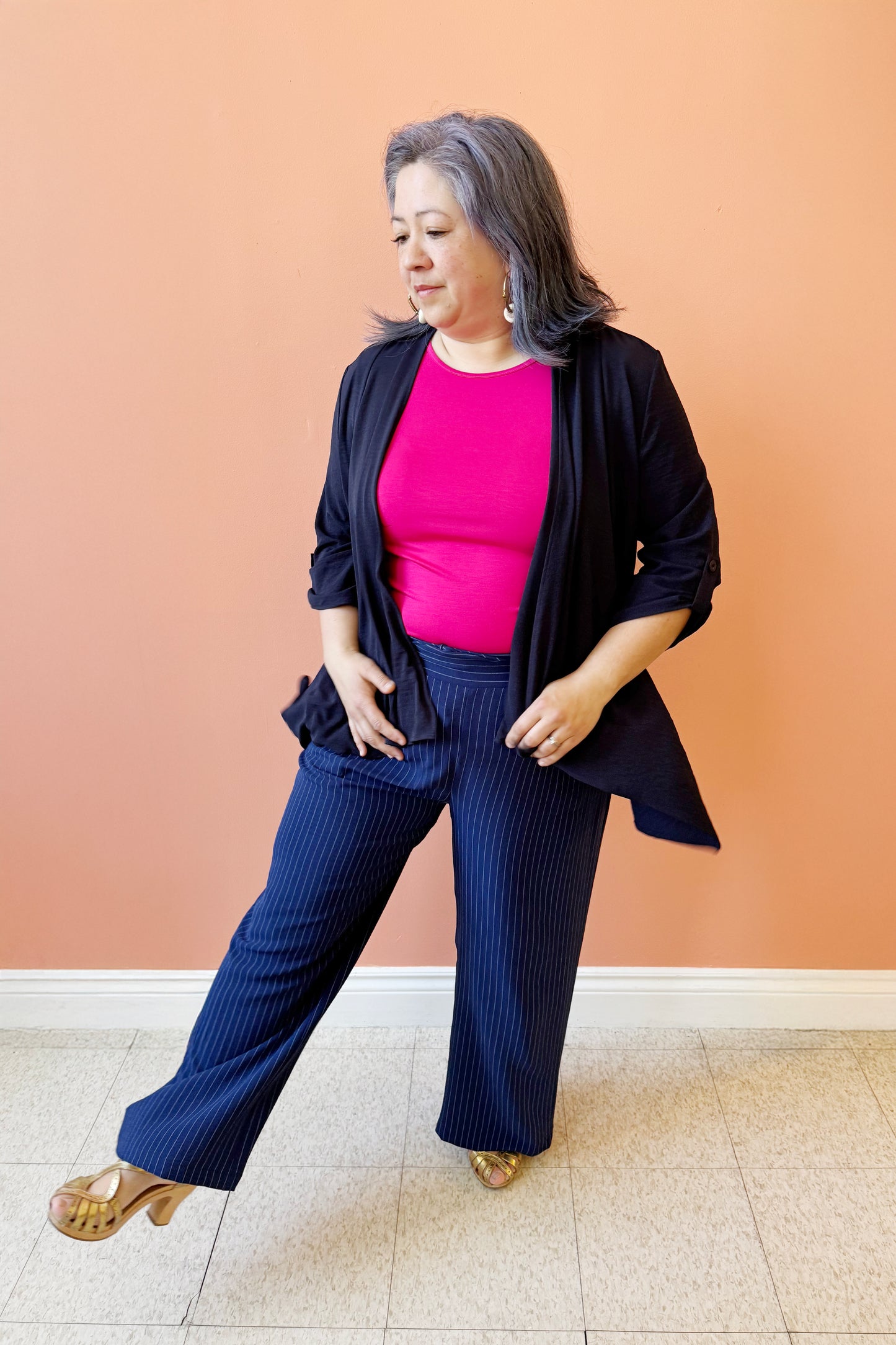 Christina is wearing the Oscar Woven Cover Up by Compli K, an open front piece with an asymmetrical front hem, sleeves that can be rolled and fastened with a button and tab, and a buckle detail at the back. She is wearing it with the Evie T-shirt in Tulip Pink and Eloise Pant in Ink pinstripe by Pure. She is standing in front of a coral wall.