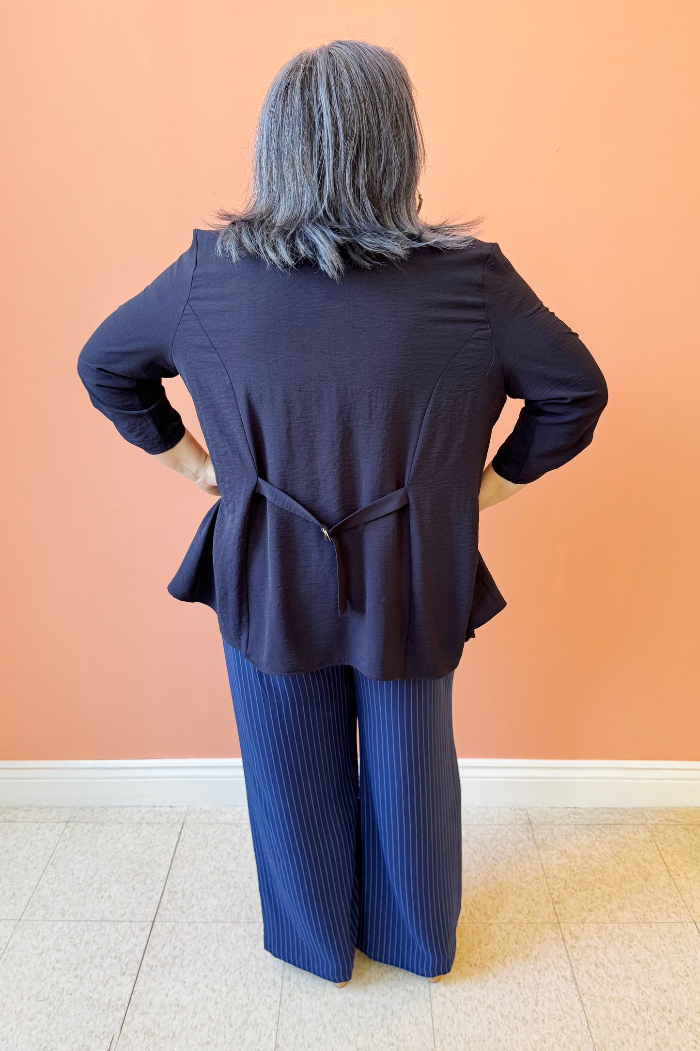 Back view of Christina wearing the Oscar Woven Cover Up by Compli K, an open front piece with an asymmetrical front hem, sleeves that can be rolled and fastened with a button and tab, and a buckle detail at the back. She is wearing it with the Evie T-shirt in Tulip Pink and Eloise Pant in Ink pinstripe by Pure. She is standing in front of a coral wall.