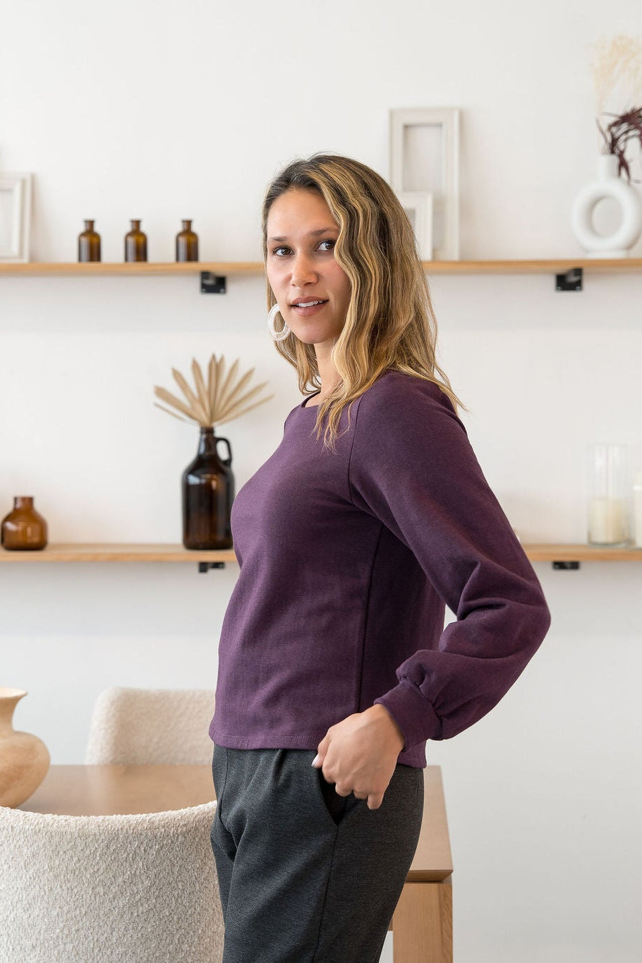 A woman wearing the Cosmopolitain Sweater by Cherry Bobin in Mauve, with a rounded neck and long sleeves with a puff detail. She is wearing it with black pants and standing in a room with shelves.