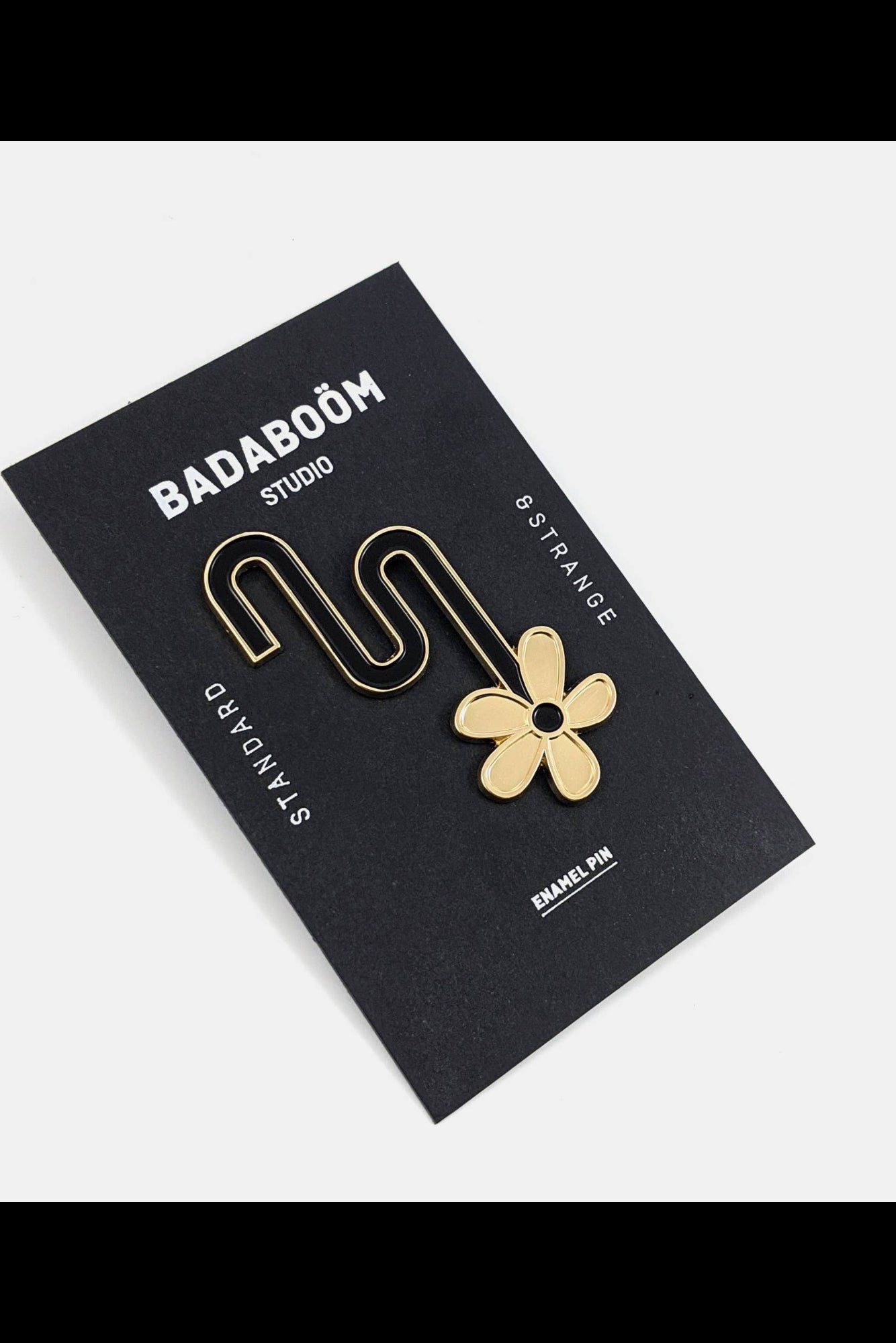 The Flower Pin from Badaboöm Studio featuring a golden flower sliding down a curved black line, shown on a black card against a white background.