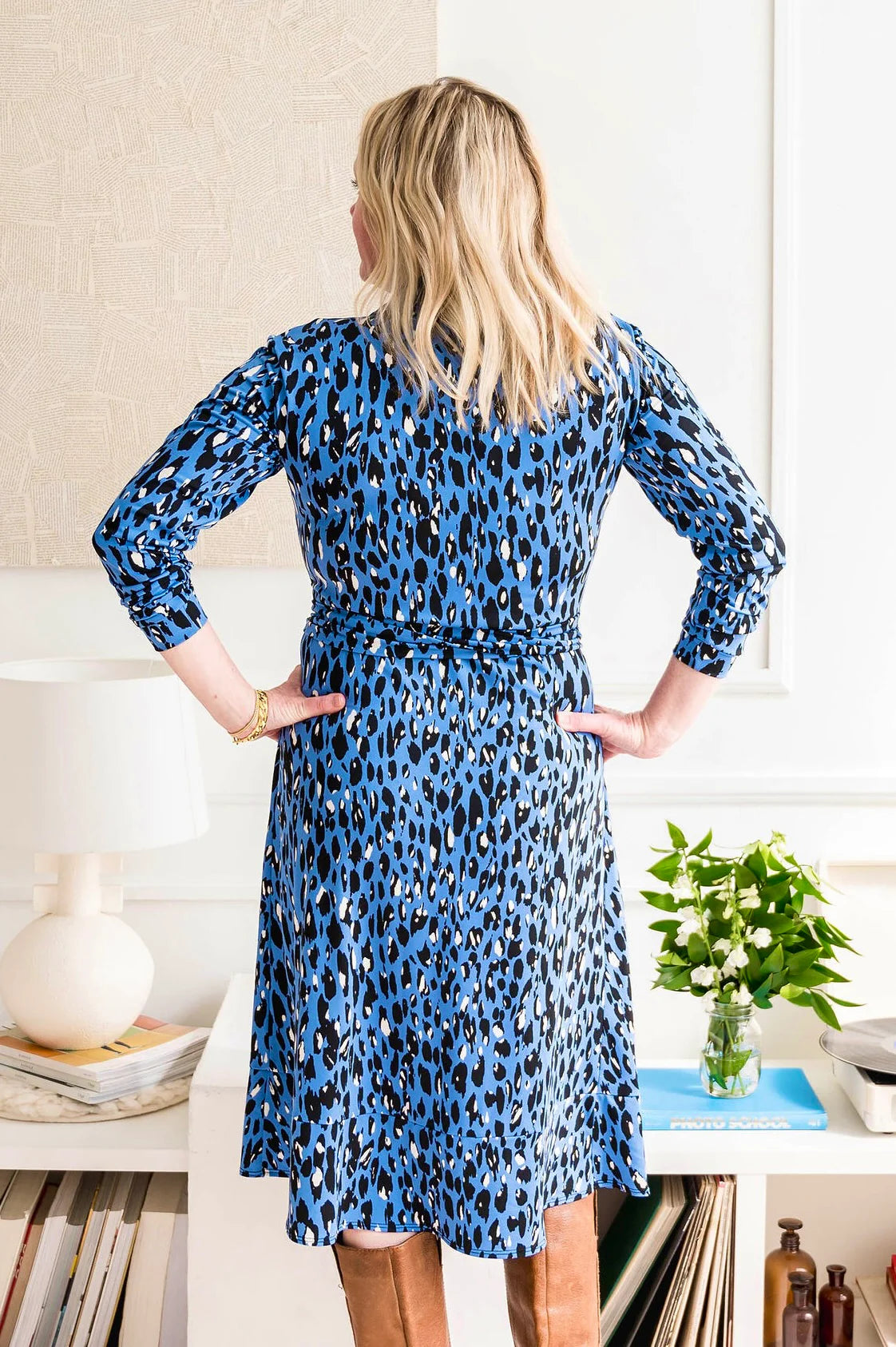Back view of a woman wearing the Leopard Wrap Dress by Dotty in Blue Leopard, a classic wrap dress with a shirt collar, adjustable waist ties, knee-length A-line skirt with a band at the hem, and long sleeves. She is standing in front of a white wall.