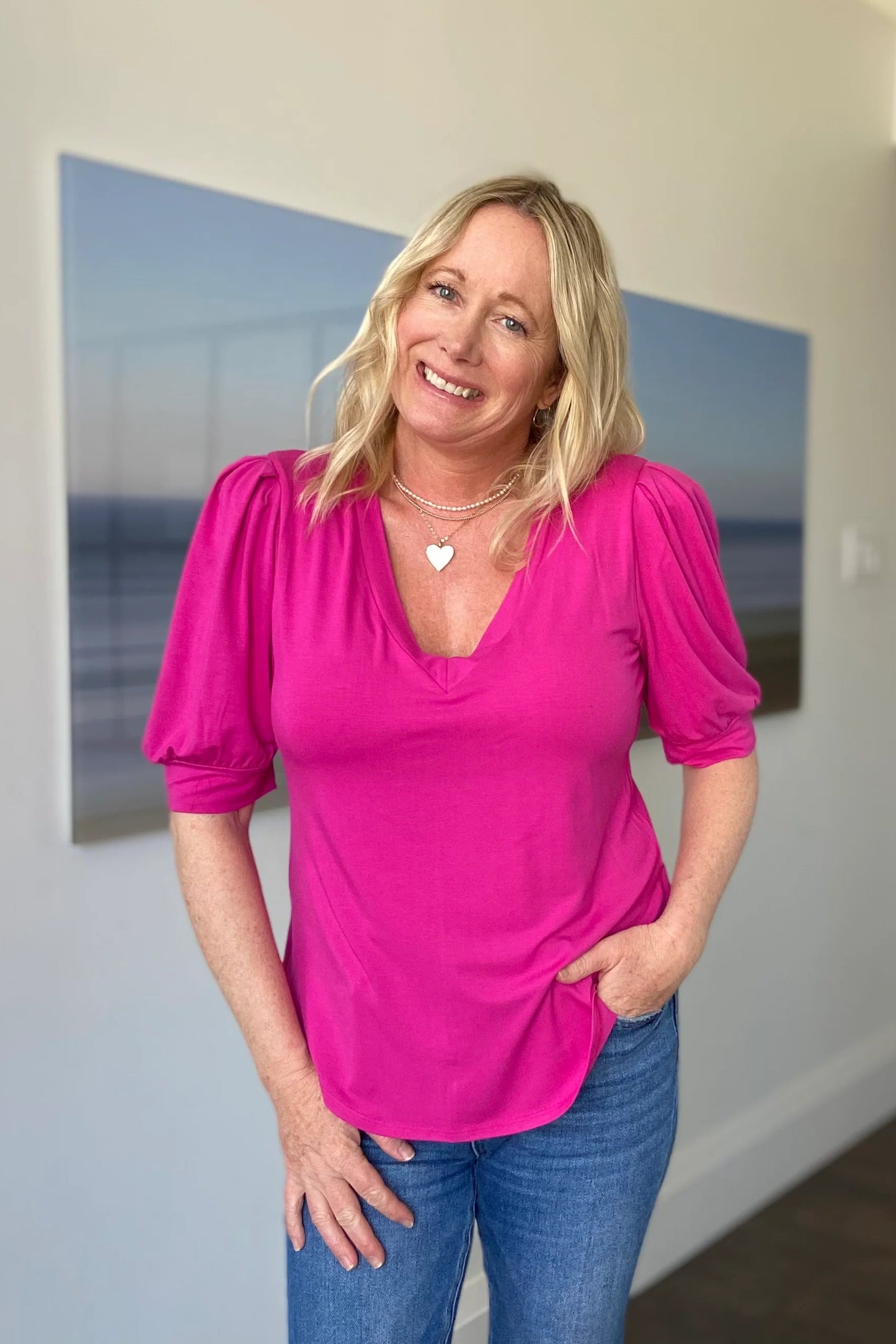 A woman wearing the Darling Tee by Dotty in Hot Pink, with a v-neck, pleats at the shoulders, elbow-length sleeves, and a rounded hem. She is wearing it with jeans and is standing in front of a white wall.