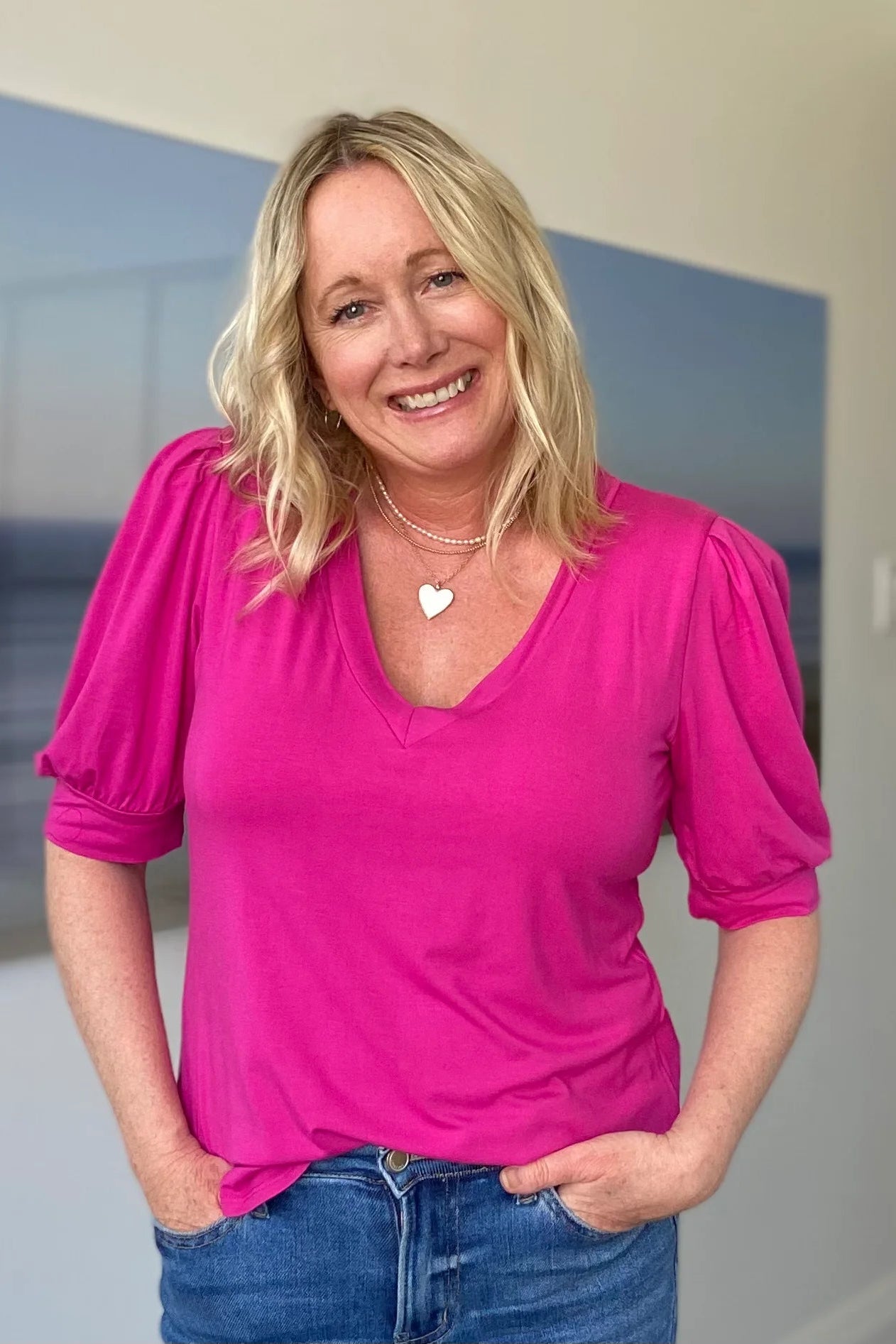 A woman wearing the Darling Tee by Dotty in Hot Pink, with a v-neck, pleats at the shoulders, elbow-length sleeves, and a rounded hem. She is wearing it with jeans and is standing in front of a white wall.