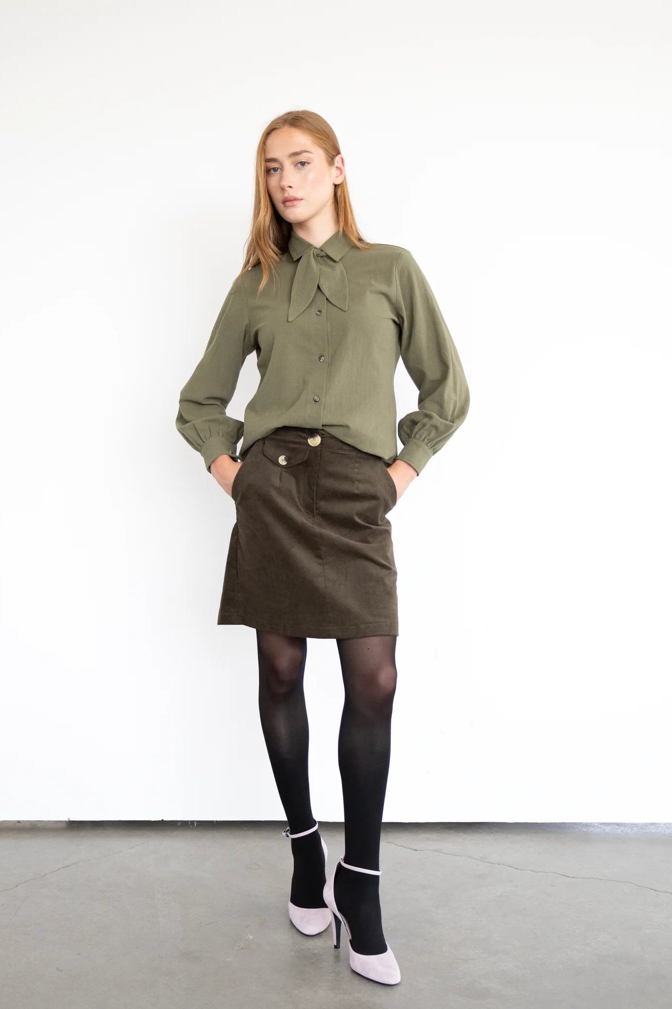 A woman wearing the Blair Shirt by Eve Lavoie in Olive, a button front shirt with a removable neck tie, rounded hem, and puffed sleeves. She is wearing it with a black skirt and standing in front of a white background.