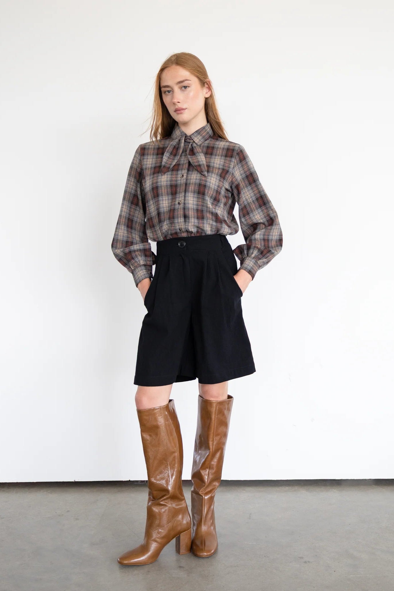 A woman wearing the Blair Shirt by Eve Lavoie in Plaid, a button front shirt with a removable neck tie, rounded hem, and puffed sleeves. She is wearing it with a black skirt and standing in front of a white background.