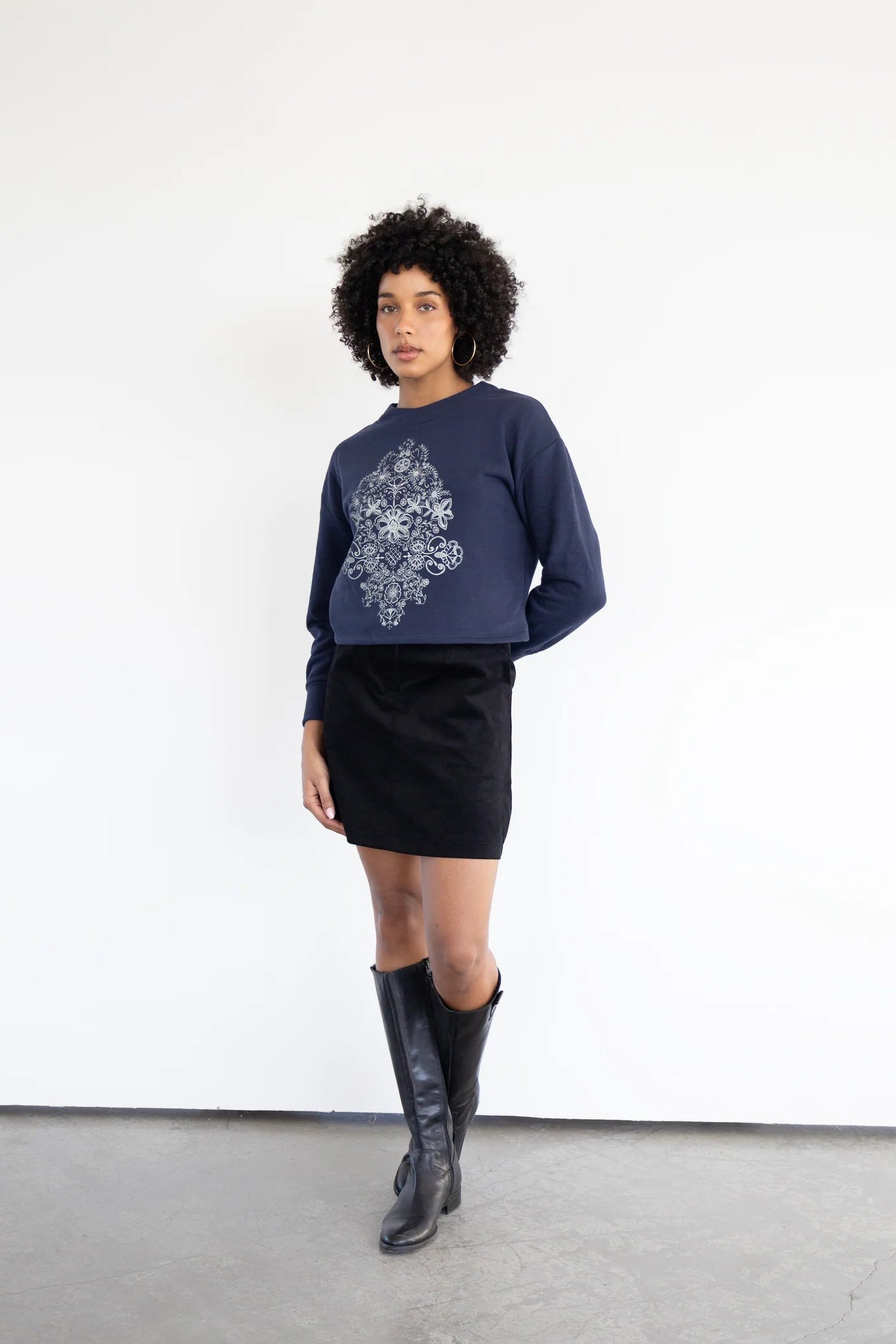 A woman wearing the Hanay Sweatshirt by Eve Lavoie in Navy Blue, a slightly cropped sweatshirt with a hand-printed silkscreen design. she is wearing it with a black skirt and standing in front of a white background.