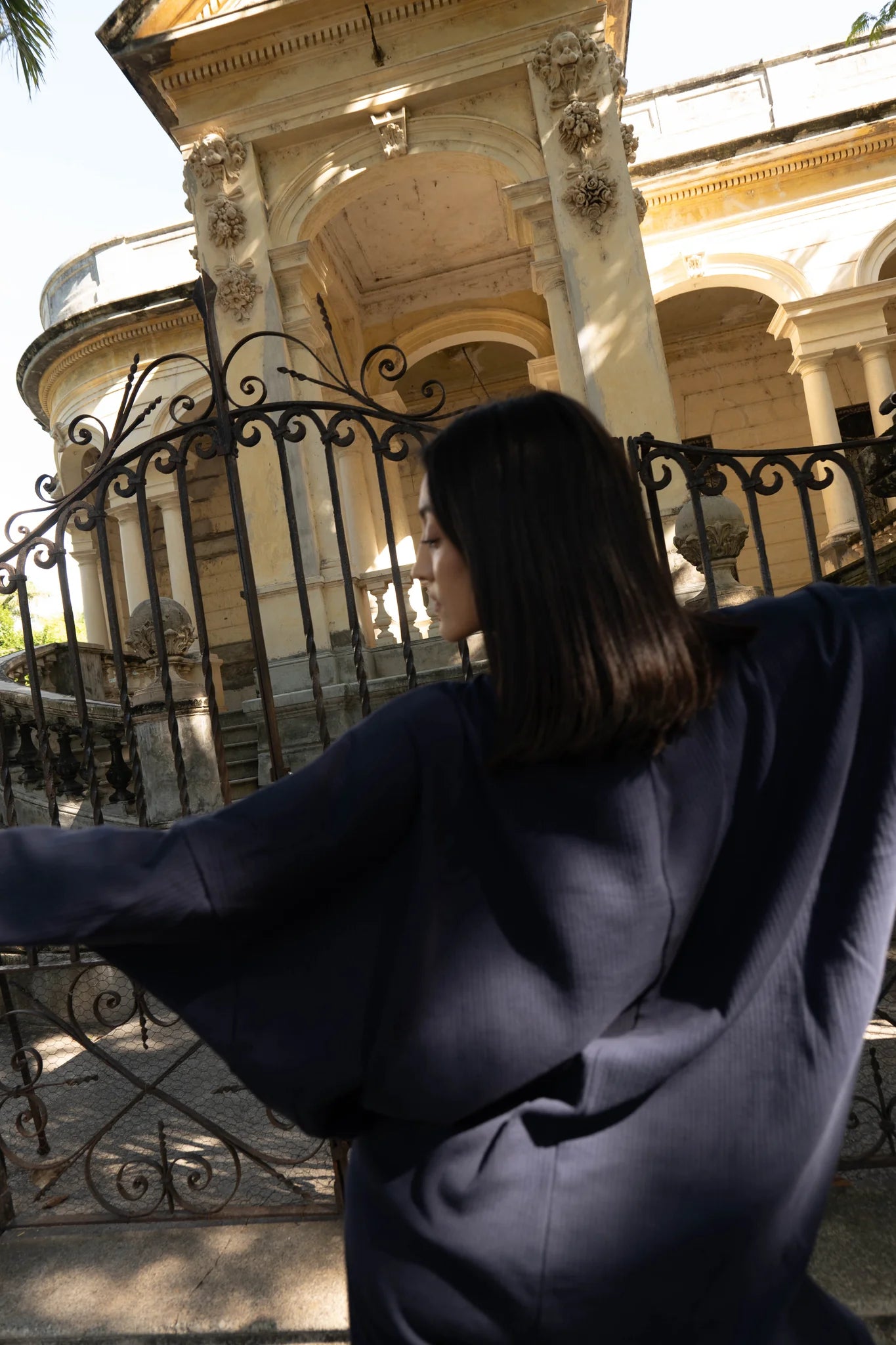 Back view of a woman wearing the Frankfurt Dress by MAS in Blueberry, a One Size ribbed cotton dress with a mock turtleneck, long batwing sleeves, and midi length. She is standing outside a historic building.