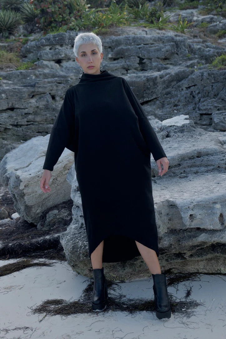 Back view of a woman wearing the Frankfurt Dress by MAS in Pepper, a One Size ribbed cotton dress with a mock turtleneck, long batwing sleeves, and midi length. She is standing on a rocky beach.
