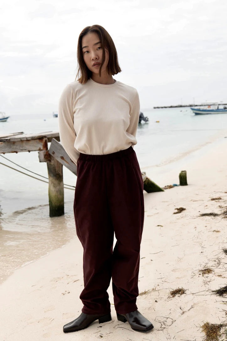 A woman wearing the Funchal Pants by MAS in Fig, fine wale corduroy pants with an elastic waist and straight cut legs. She is wearing them with a white top and is standing on a beach.