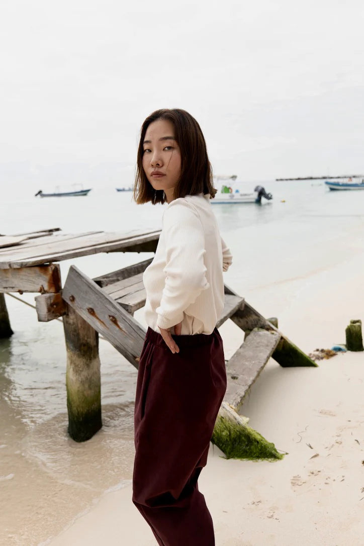 Sid view of a woman wearing the Whistler Long Sleeve Top in Almond, a ribbed cotton knit with a round neck and straight cut. She is wearing it with the Funchal Pants in Fig and standing on a beach.