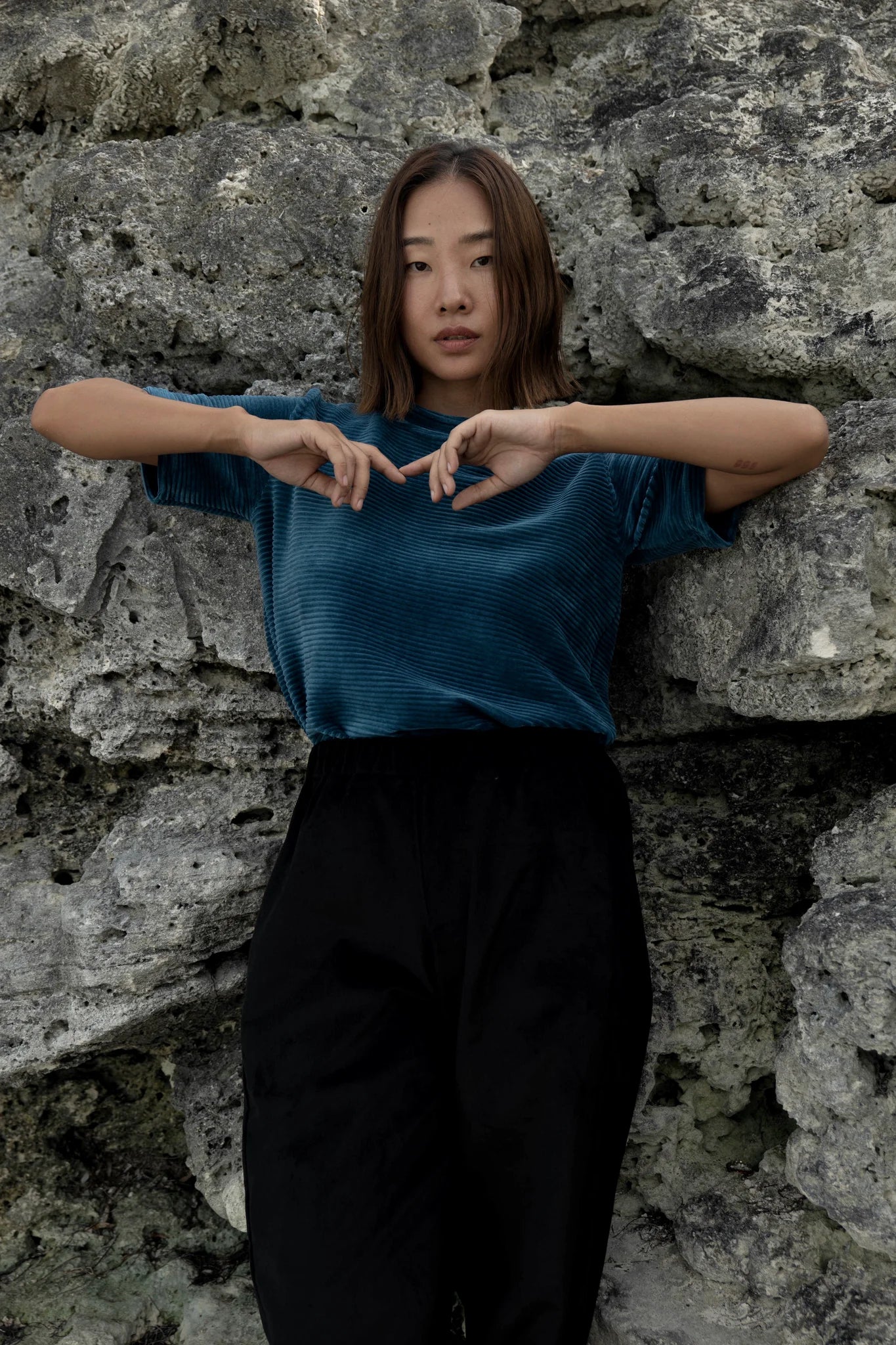 A woman wearing the Tofino T-Shirt by MAS in Juniper, a short sleeved top in ribbed velvet. She is wearing it with the Funchal Pants in Pepper and standing against a rock wall.