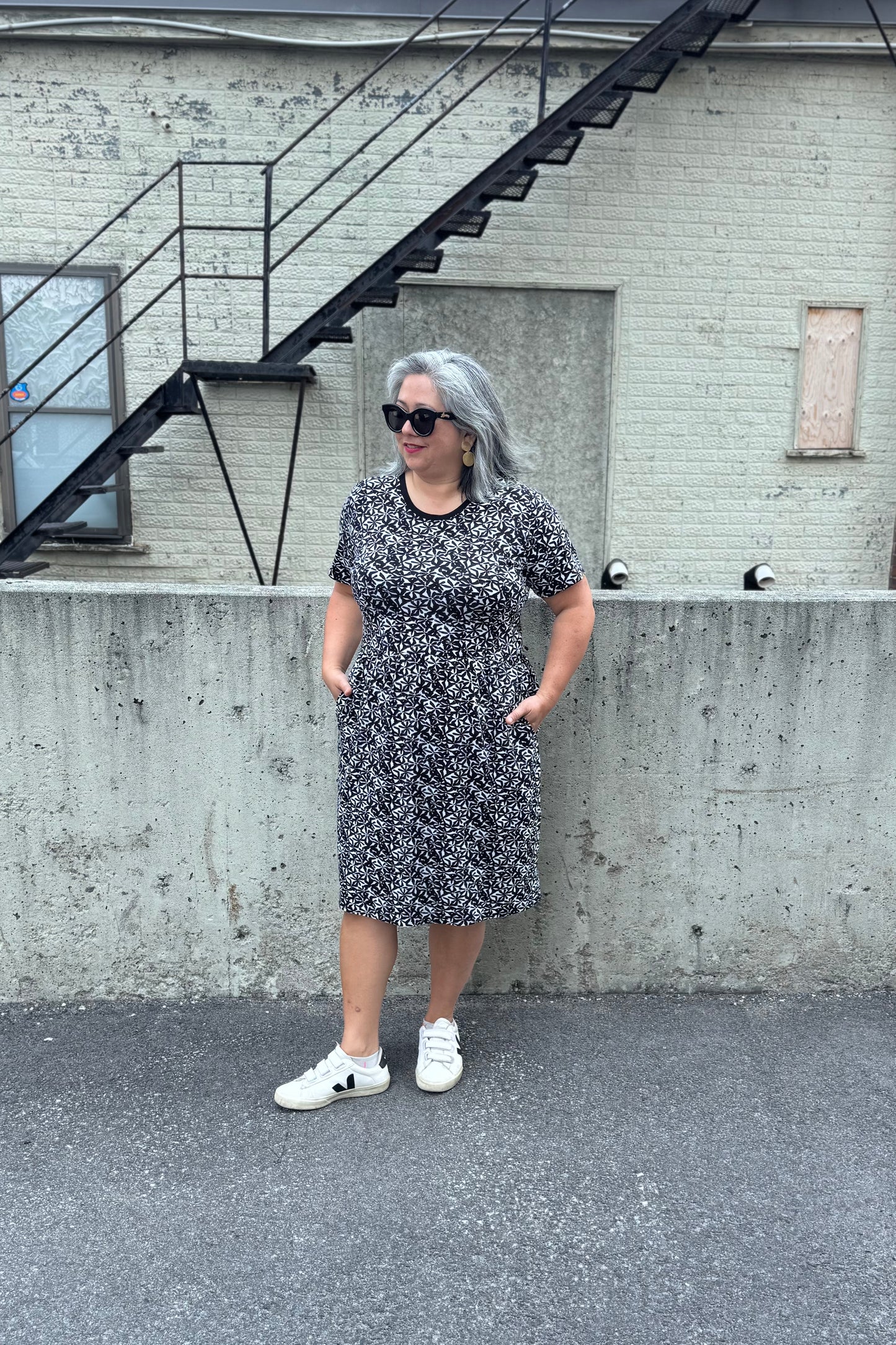 A woman wearing the Hayley Dress by Tangente in Black and White floral print, a short sleeved bamboo dress with a gathered knee-length skirt. She is standing outside in front of a cement wall.