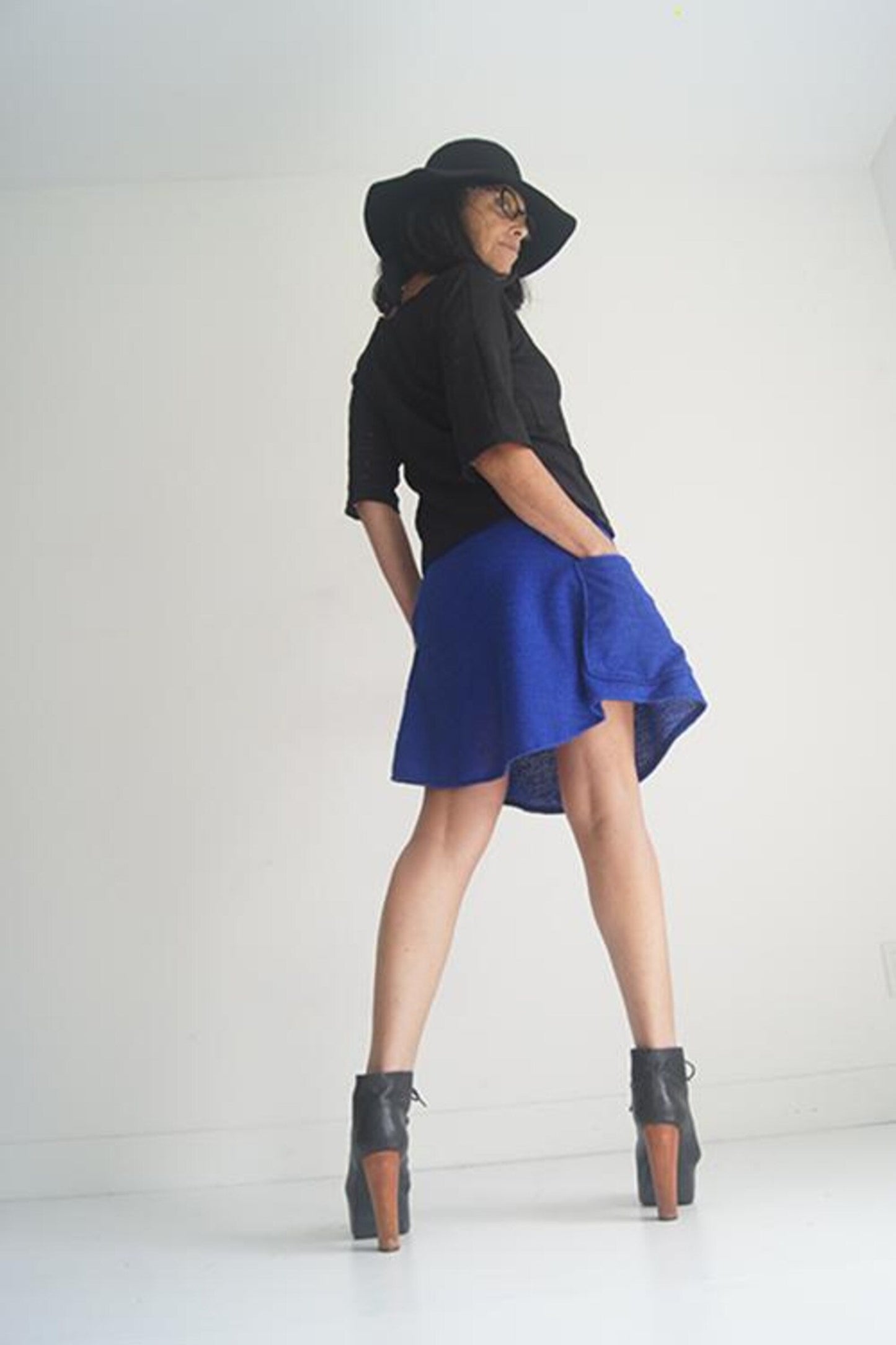 Side view of a woman wearing the Short Skirt by SI Design in Blue, an above-the-knee A-line skirt with an elastic waist and patch pockets. She is wearing it with a black top and standing in front of a grey background.