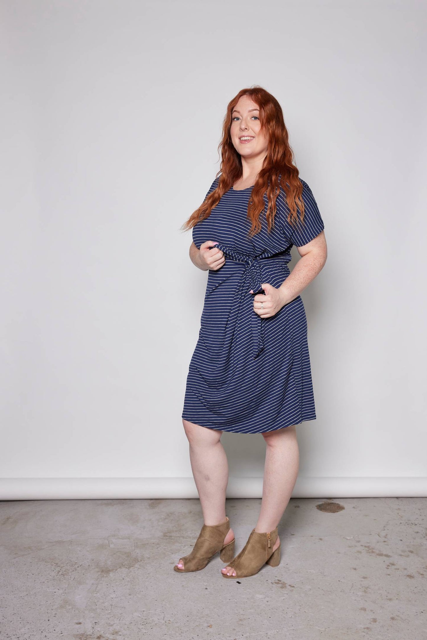 A woman wearing the Joelle Dress by Tangente in Blue/Grey, a cap-sleeved dress in striped bamboo with a tie at the waist and an above-the-knee hem. She is standing in front of a white wall.