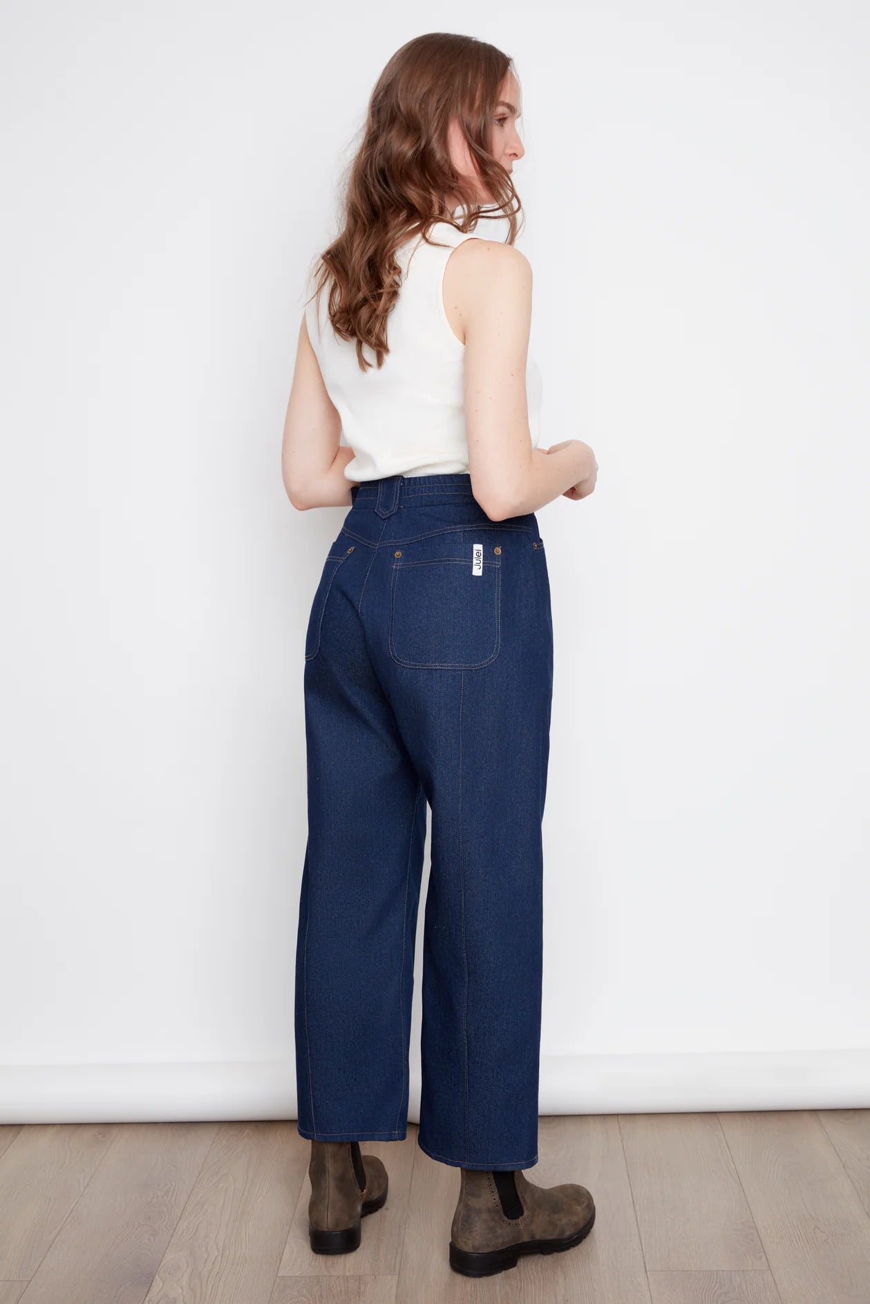 Bak view of a woman wearing the Alice Straight Leg Jeans by Julei in Dark Denim, with an elastic back waistband, seams at the front and back of the legs, and pointed belt loops. She is wearing them with a white tank and standing in front of a white background.