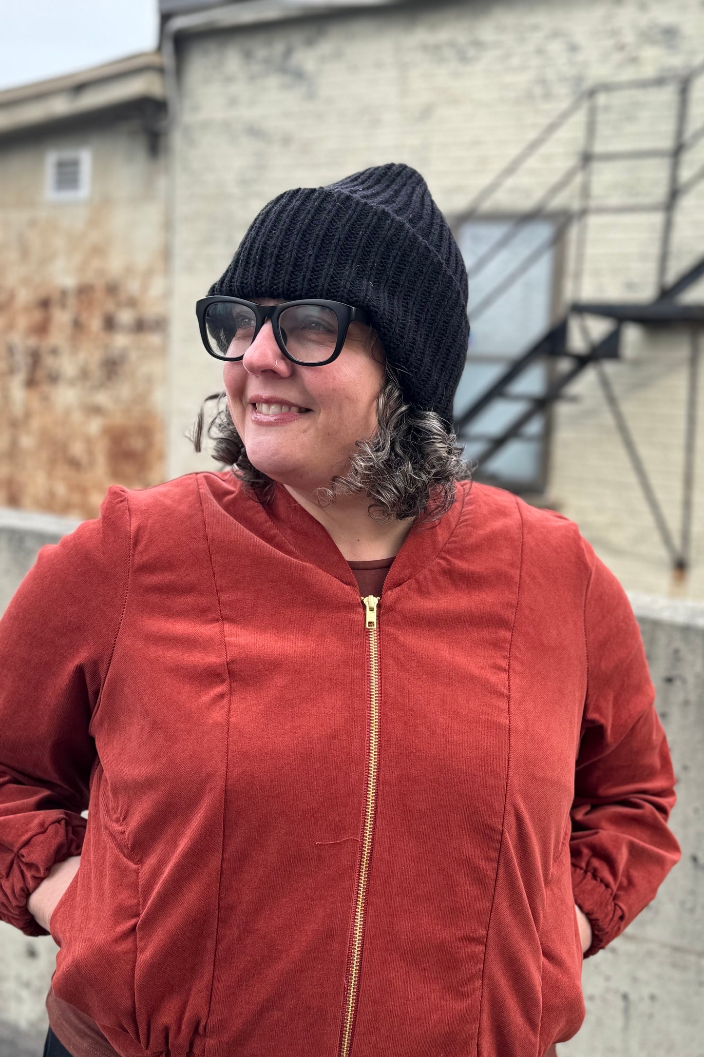 A woman wearing the Ribbed Beanie by Charlevoix Pure Laine in Black. She is wearing it with the Redrawn Zip Jacket by Eve Lavoie and is standing outside.