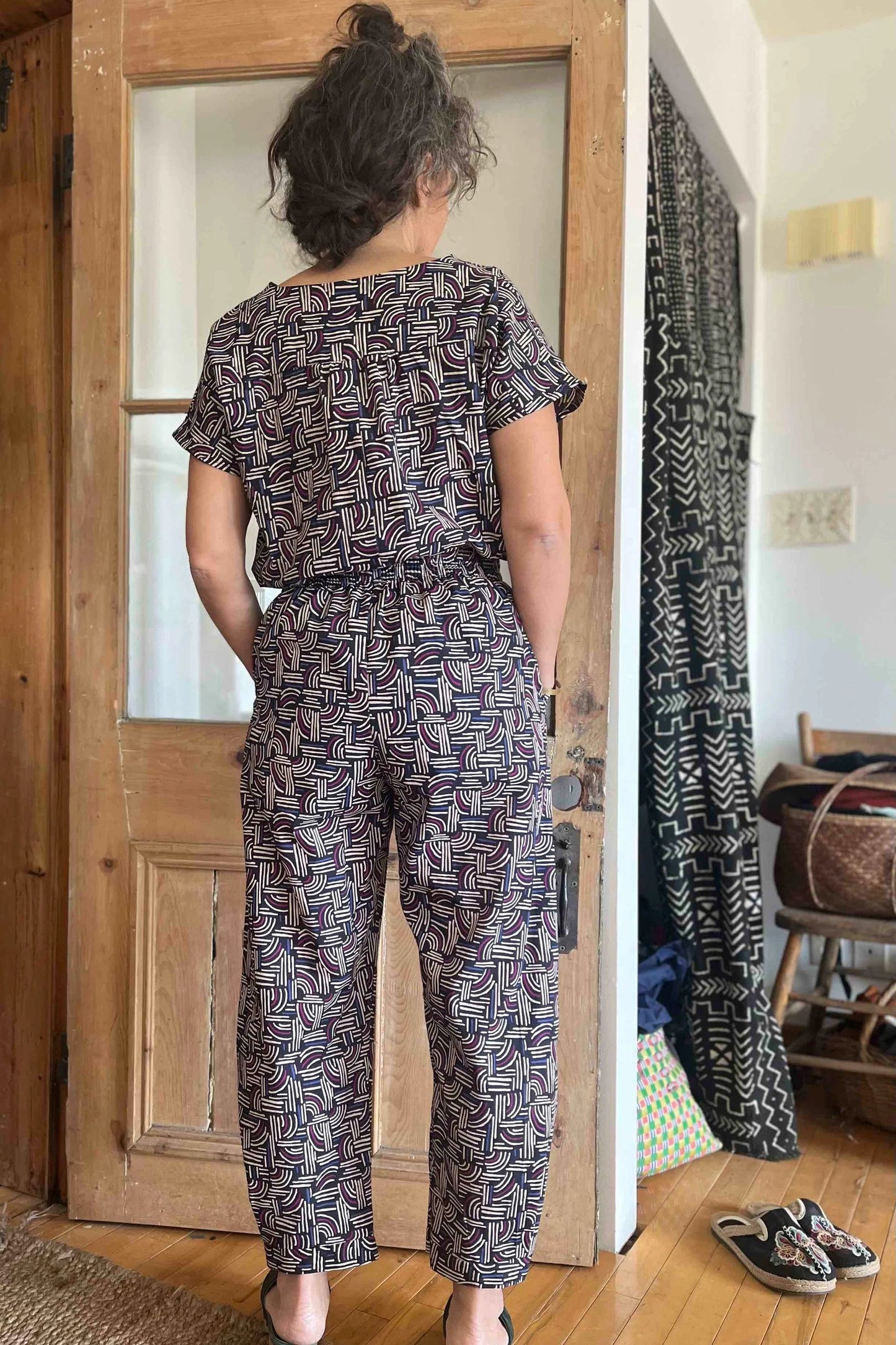 Back view of a woman wearing the Sienna Pants by Kazak in Night Basketwork print, featuring a flat front waistband with elastic at the sides and back, large angled pockets, and loose fitting legs that taper slightly at the ankle. She is wearing them with the matching Lucca Top and is standing in front of a wooden door.