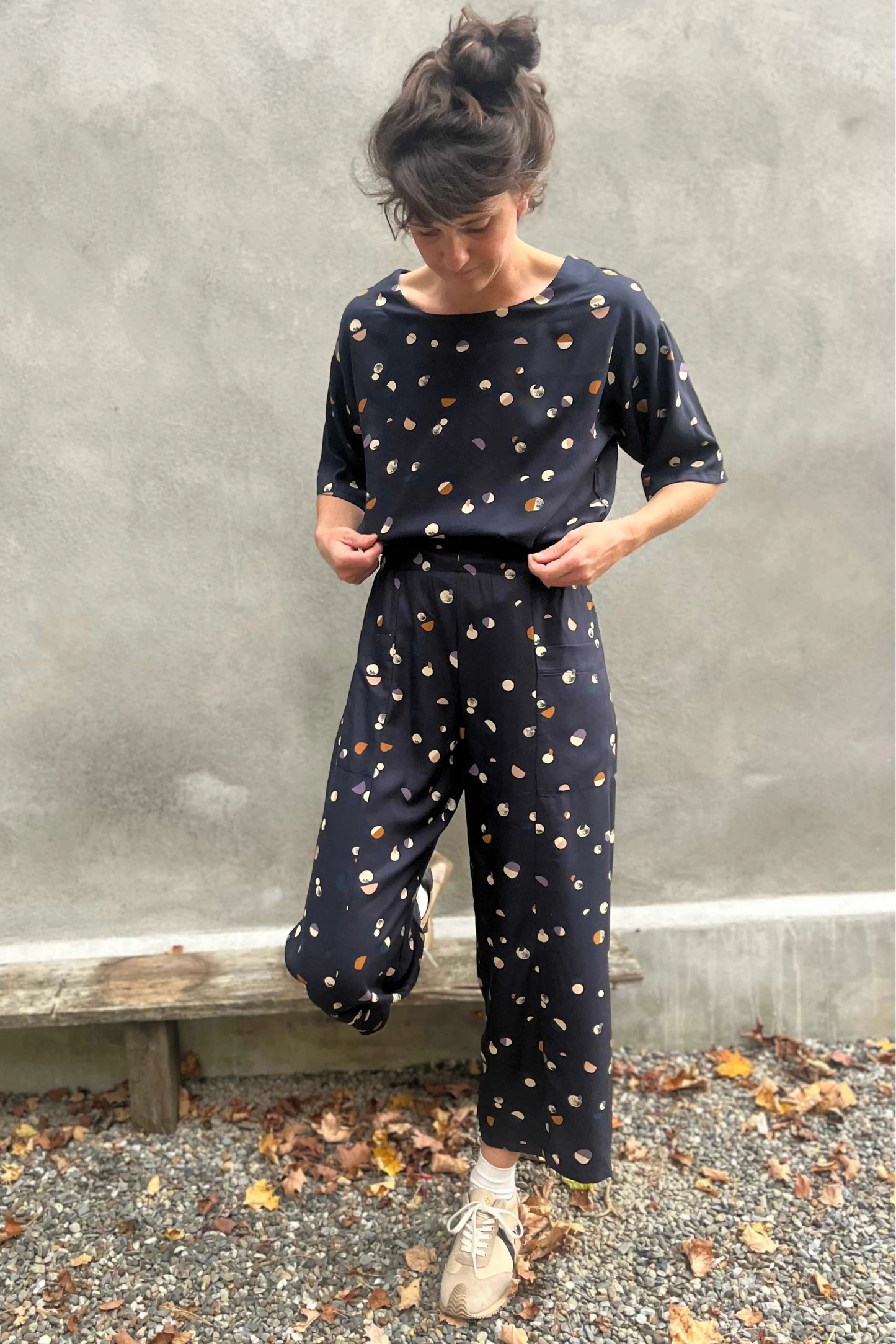 A woman wearing the Olea Pants by
Kazak in Midnight Moons, wide-legged ankle-length pants with a flat-front elastic waist and large patch pockets. She is wearing them with the matching Abelia Top and standing outside by a grey wall.