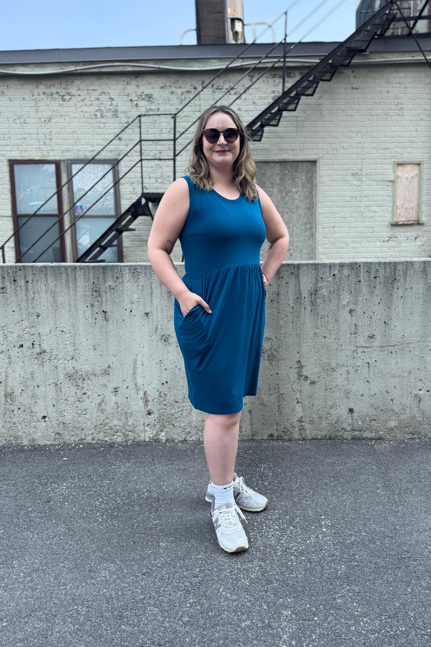 A woman wearing the Mai Dress by Tangente in Teal, a sleeveless bamboo jersey dress with a round neck and gathered knee-length skirt. She is standing outside in front of a cement wall.