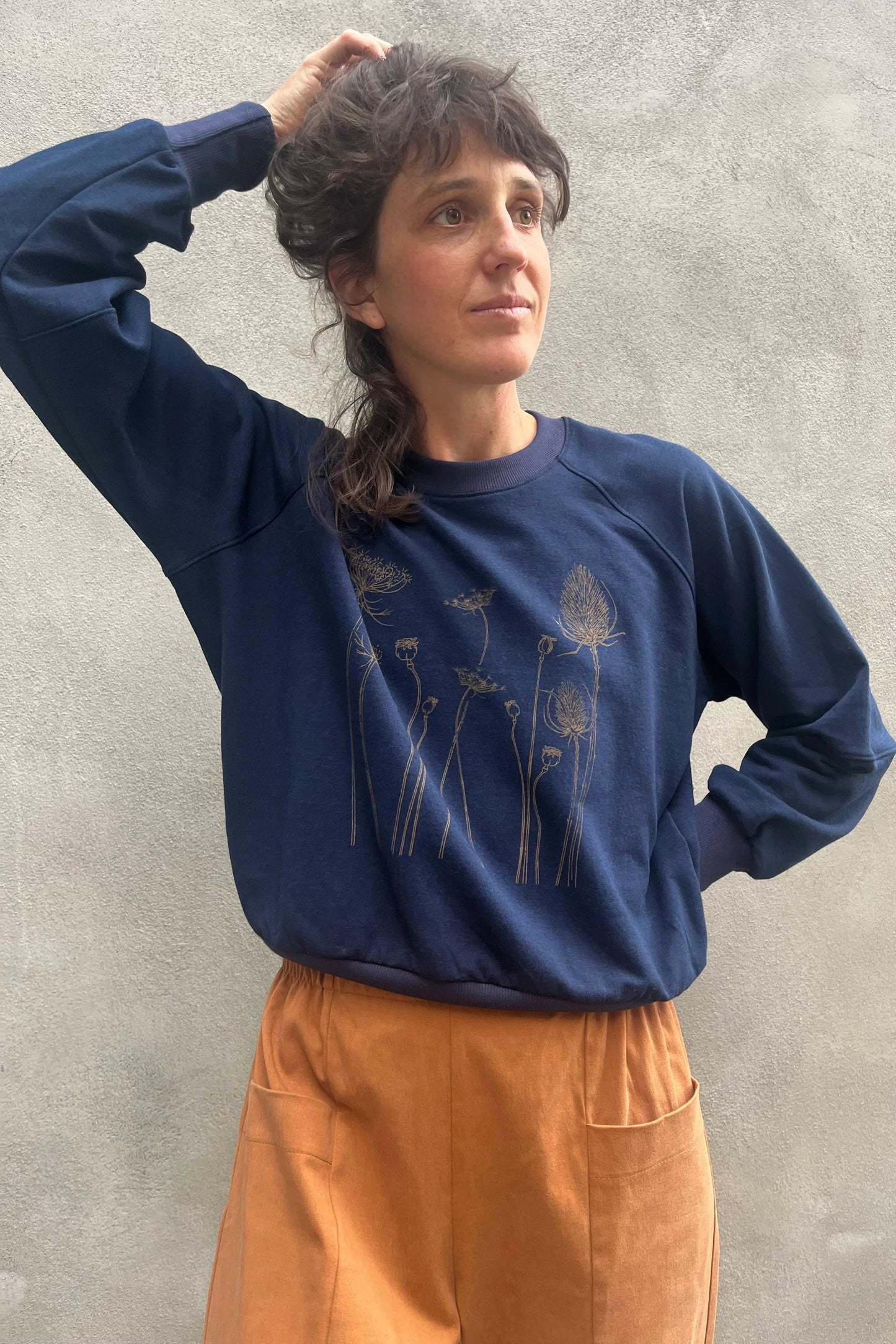 A woman wearing the Marilou Crewneck with Flowers by Kazak in Navy, a raglan sleeved sweatshirt with a screen print design. She is wearing it with yellow and standing by a grey wall.