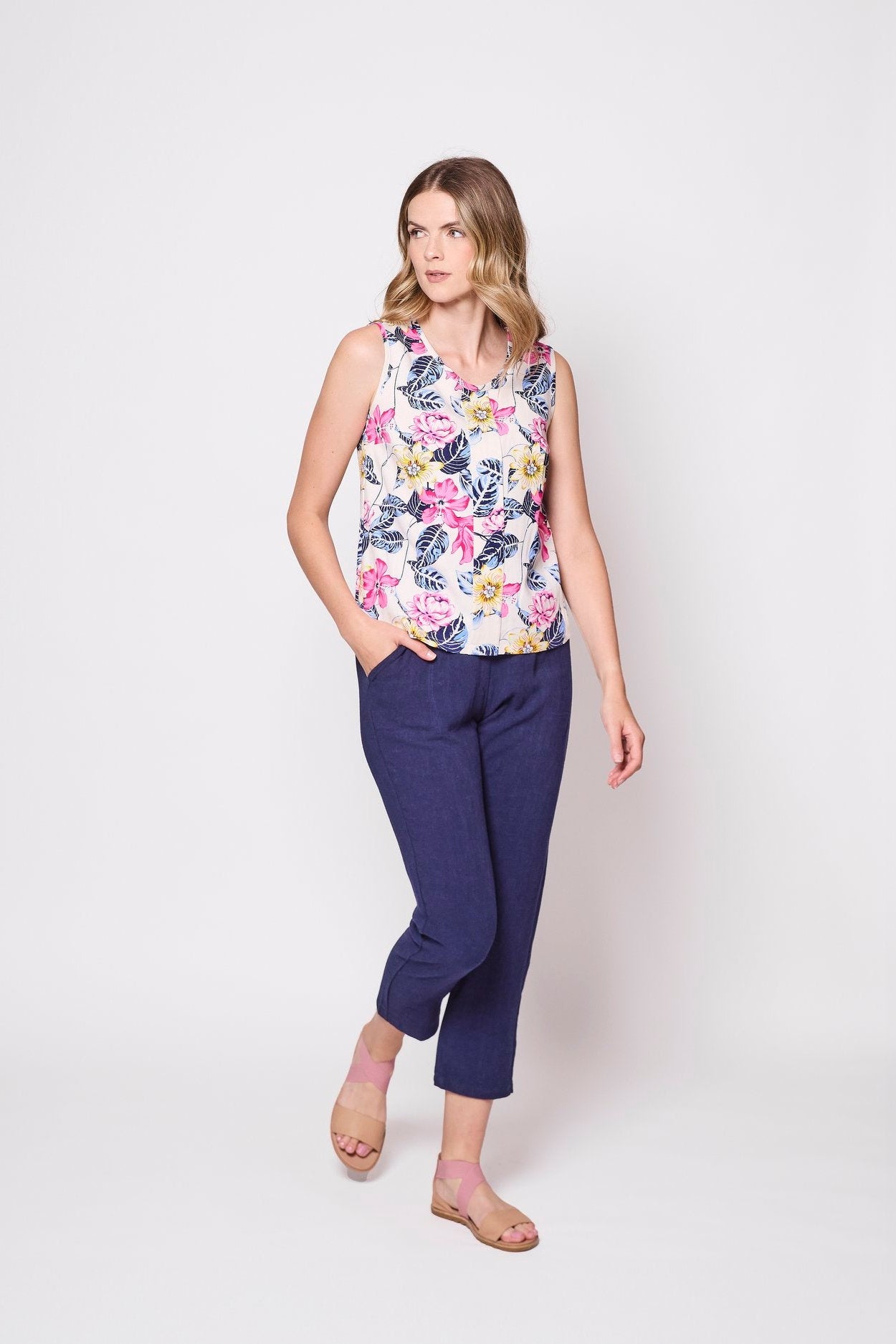 A woman wearing the Marney
Tank Top by Tangente in White Floral, with a round neck, full-length centre pleat, and darts at the bust. She is wearing it with navy pants and standing in front of a white background.