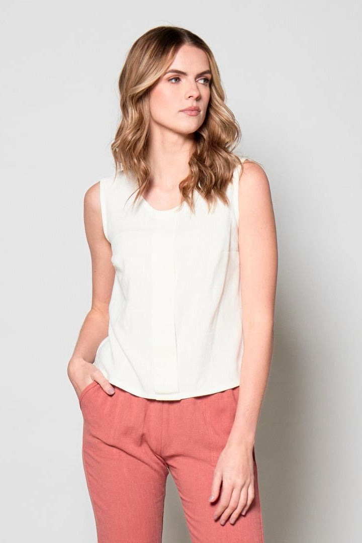 3/4 view of a woman wearing the Marney Tank Top by Tangente in White with a round neck, full-length centre pleat, and darts at the bust. She is wearing it with coral pants and standing in front of a white background.