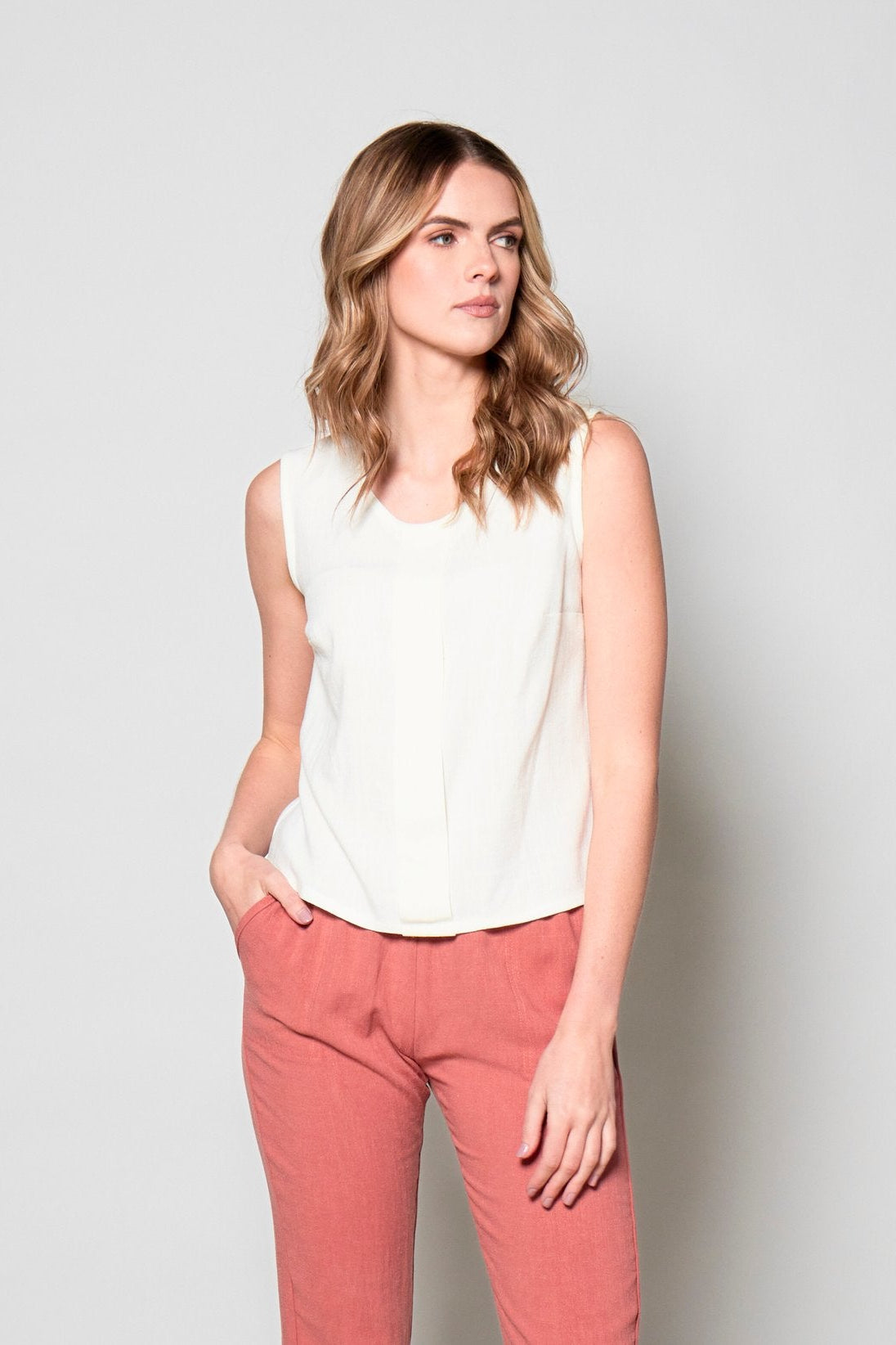 3/4 view of a woman wearing the Marney Tank Top by Tangente in White, with a round neck, full-length centre pleat, and darts at the bust. She is wearing it with coral pants and standing in front of a white background.
