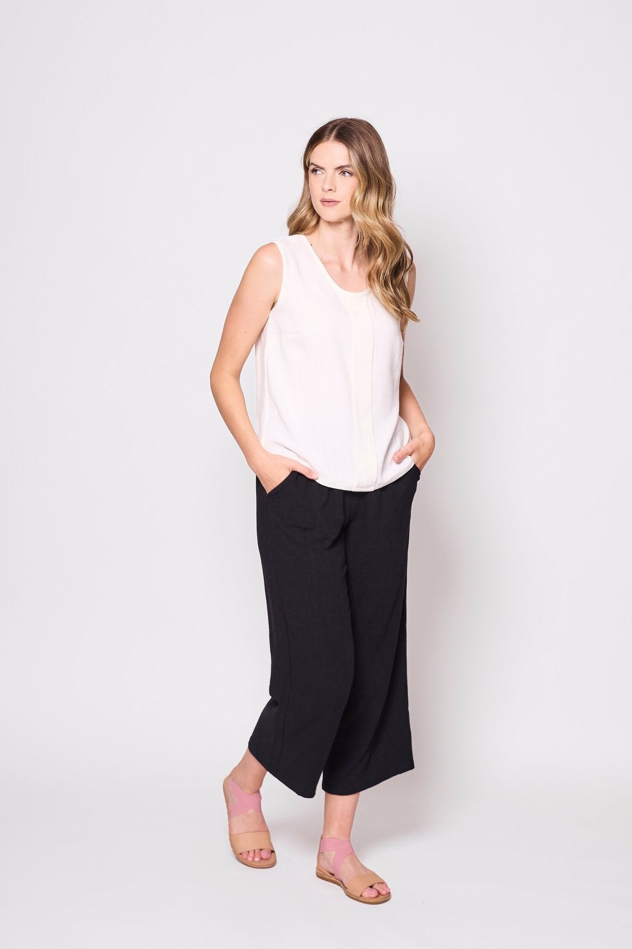 A woman wearing the Marney Tank Top by Tangente in White, with a round neck, full-length centre pleat, and darts at the bust. She is wearing it with black pants and standing in front of a white background.