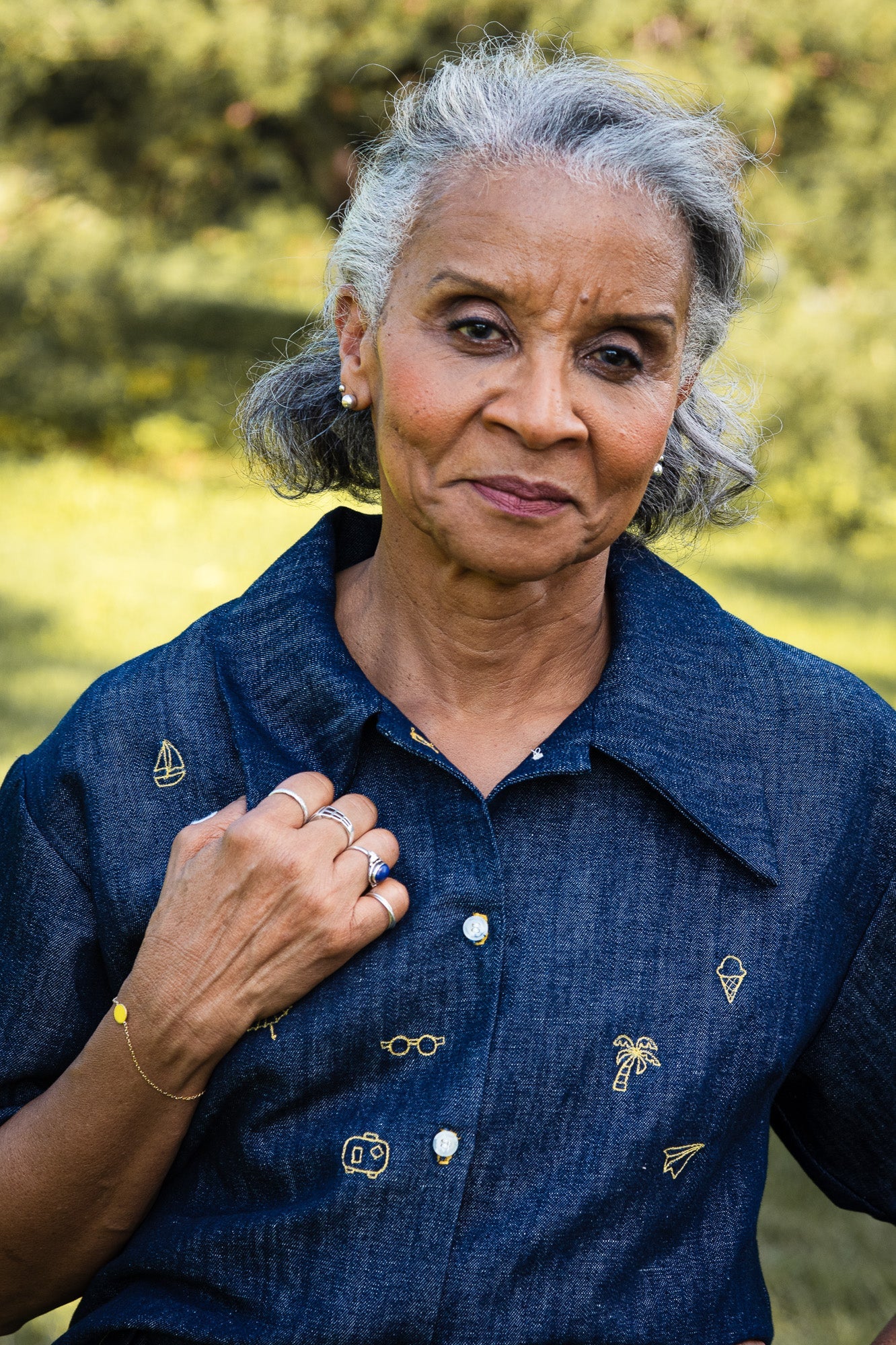 Close-up of a woman wearing the Almeria Blouse by MAS in Denim, a short-sleeved shirt with a large collar, button front, a boxy fit and hand-embroidered designs on the front. She is standing outside.