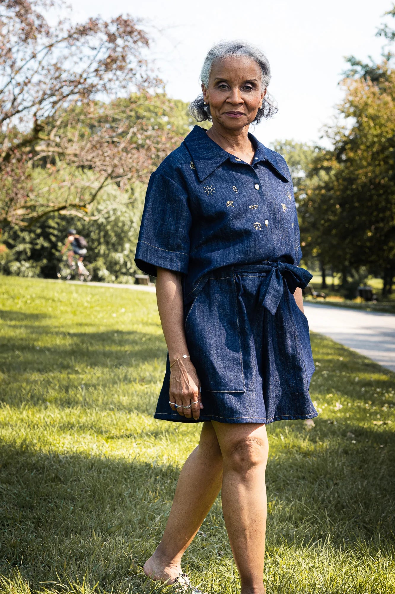 Close-up of a woman wearing the Almeria Blouse by MAS in Denim, a short-sleeved shirt with a large collar, button front, a boxy fit and hand-embroidered designs on the front. She is wearing it with the Cali Short in Denim and is standing outside.