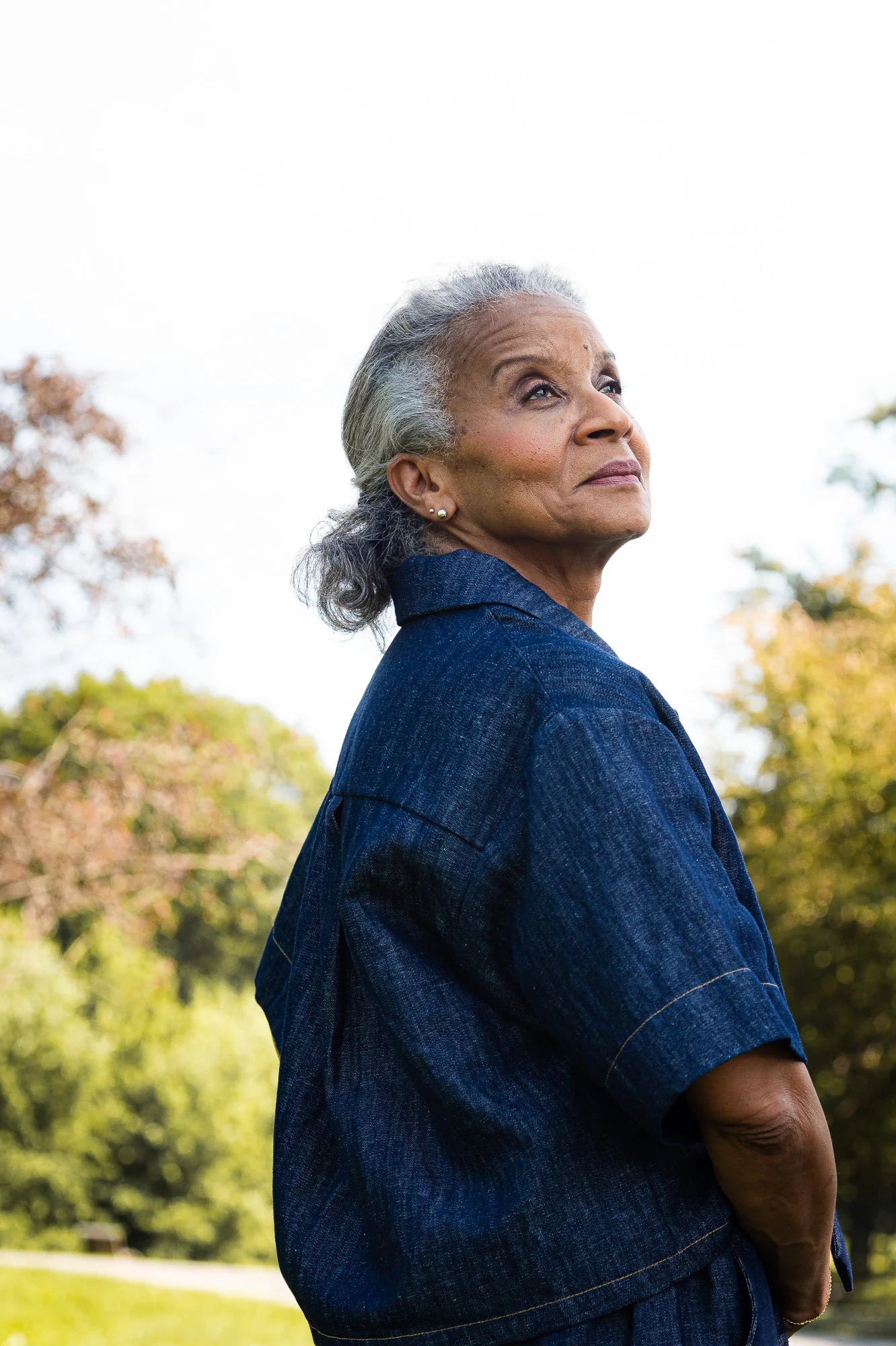 Close-up side view of a woman wearing the Almeria Blouse by MAS in Denim, a short-sleeved shirt with a large collar, button front, a boxy fit and hand-embroidered designs on the front. She is standing outside.