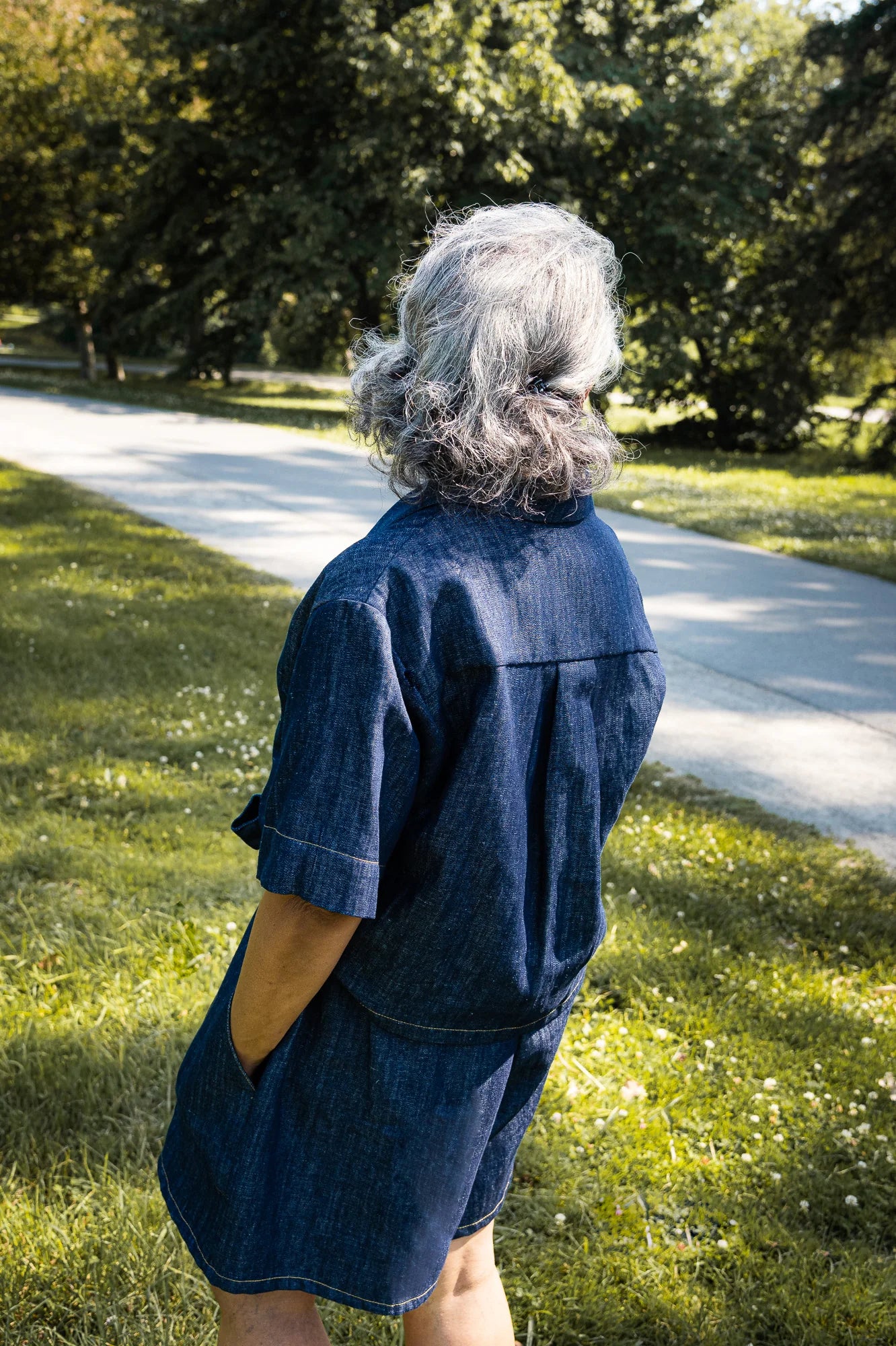 Back view of a woman wearing the Almeria Blouse by MAS in Denim, a short-sleeved shirt with a large collar, button front, a boxy fit and hand-embroidered designs on the front. She is wearing it with the Cali Short in Denim and is standing outside.