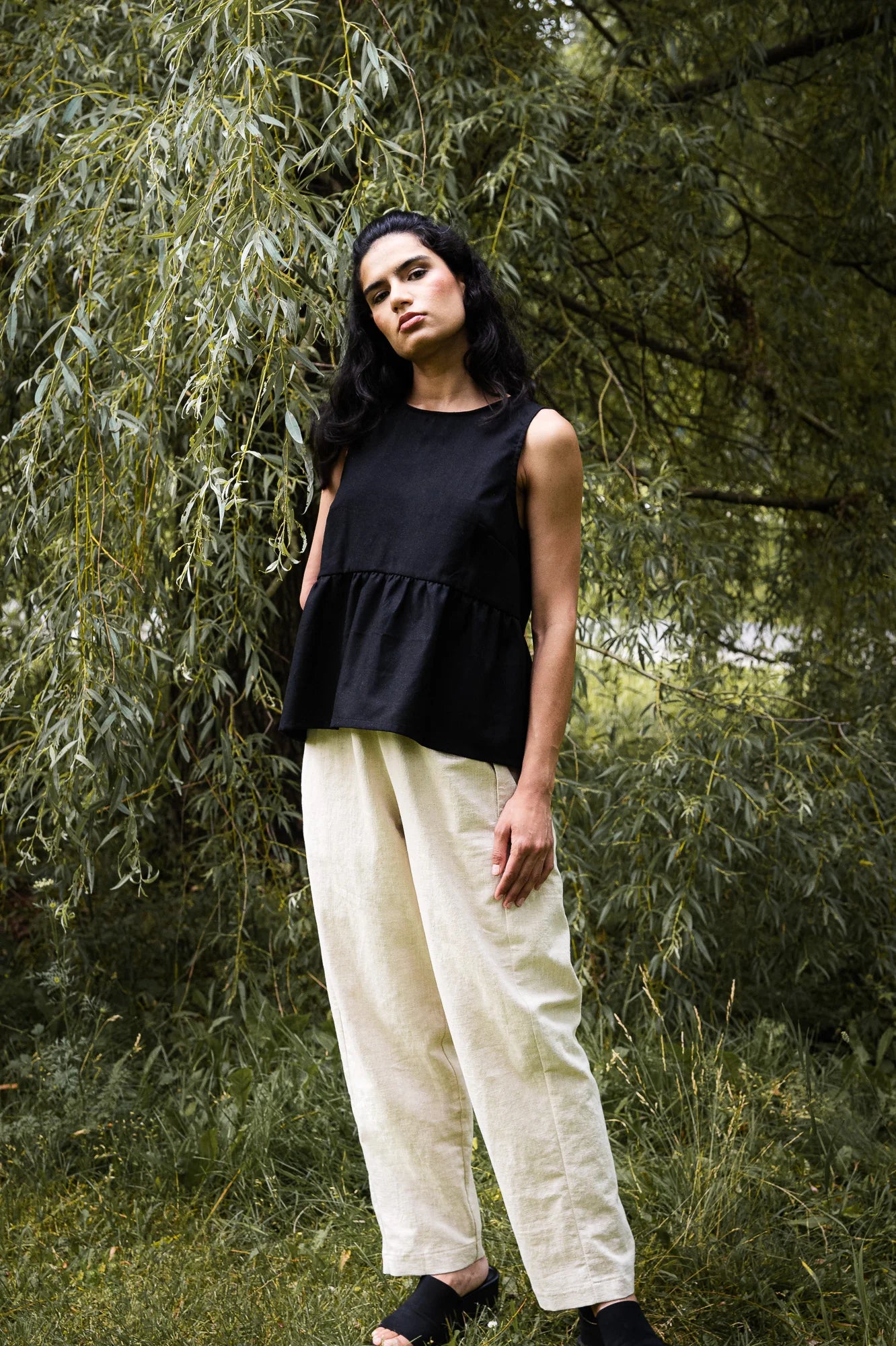A woman wearing the Malé Tank Top by MAS in Pepper, with a high neck, a curved gathered seam that creates a peplum effect, and three ties at the back. She is wearing it with ivory pants and standing outside.