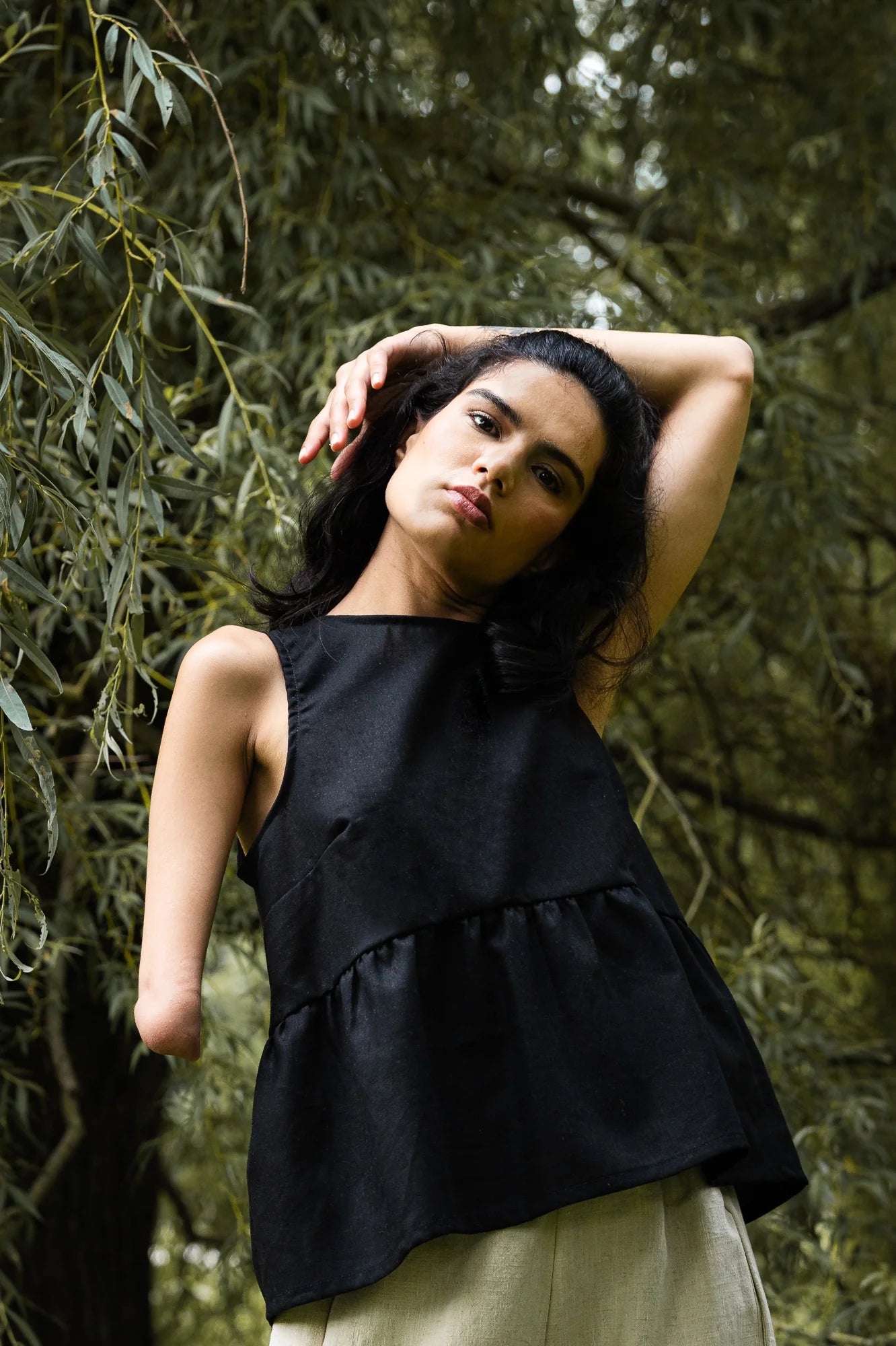 A woman wearing the Malé Tank Top by MAS in Pepper, with a high neck, a curved gathered seam that creates a peplum effect, and three ties at the back. She is standing outside. .