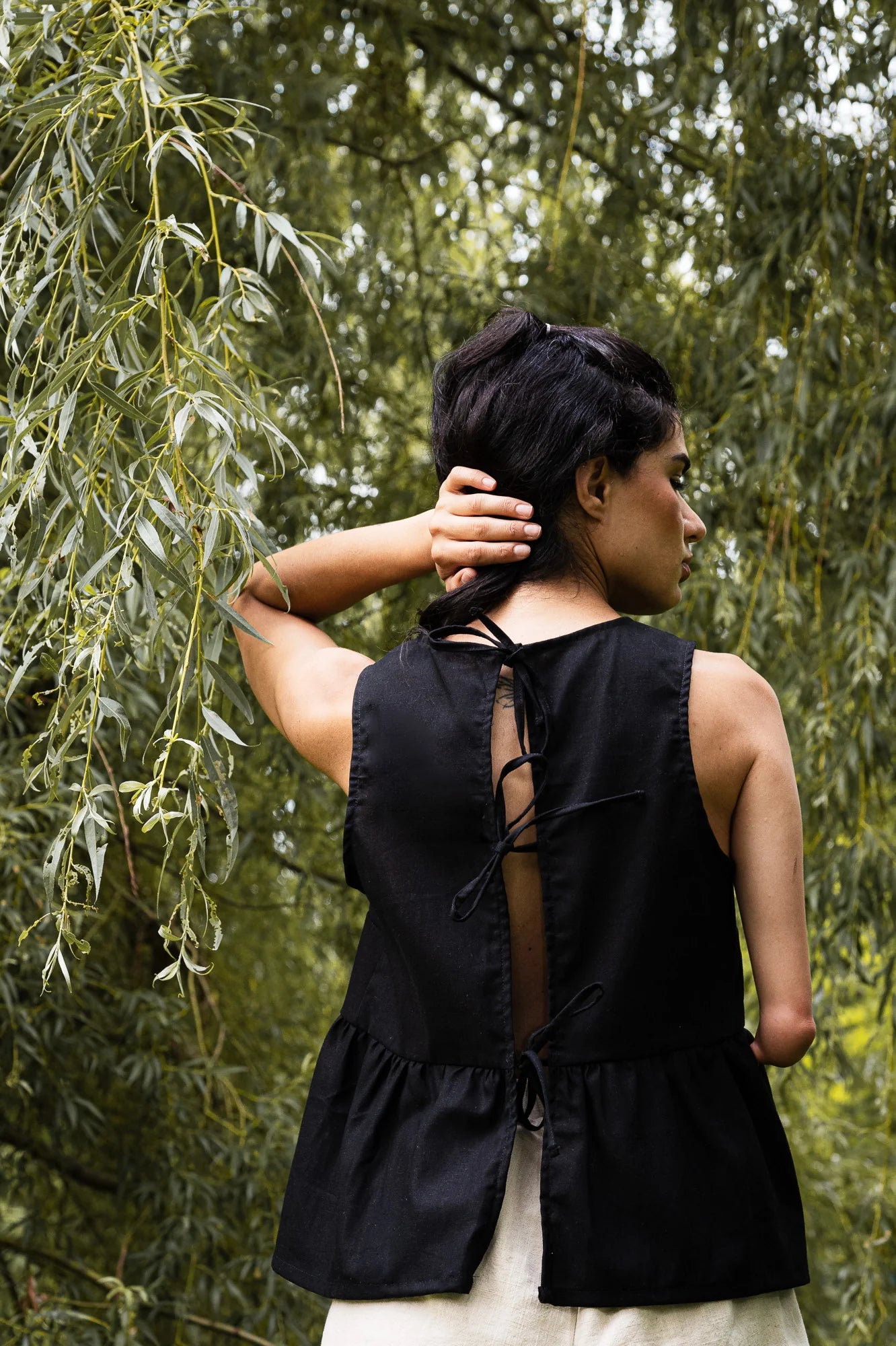 Back view of a woman wearing the Malé Tank Top by MAS in Pepper, with a high neck, a curved gathered seam that creates a peplum effect, and three ties at the back. She is standing outside. .