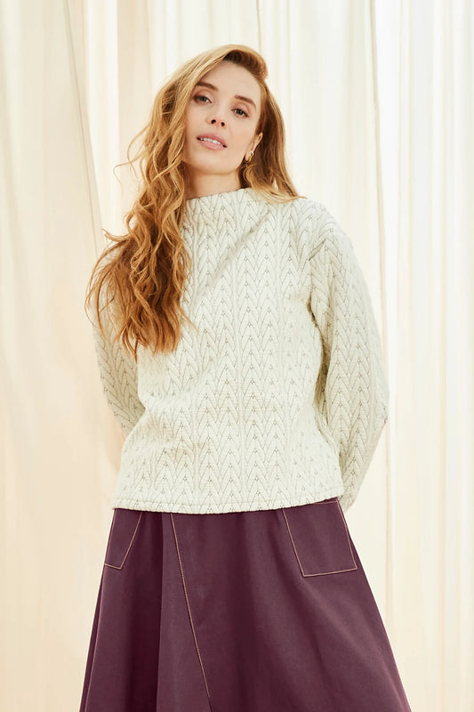 A woman wearing the Lorelai Sweater by Melow in Ecru, a quilted sweater in a cable-knit texture with a high neck and long sleeves. She is wearing it with a purple skirt and standing in front of a white background.