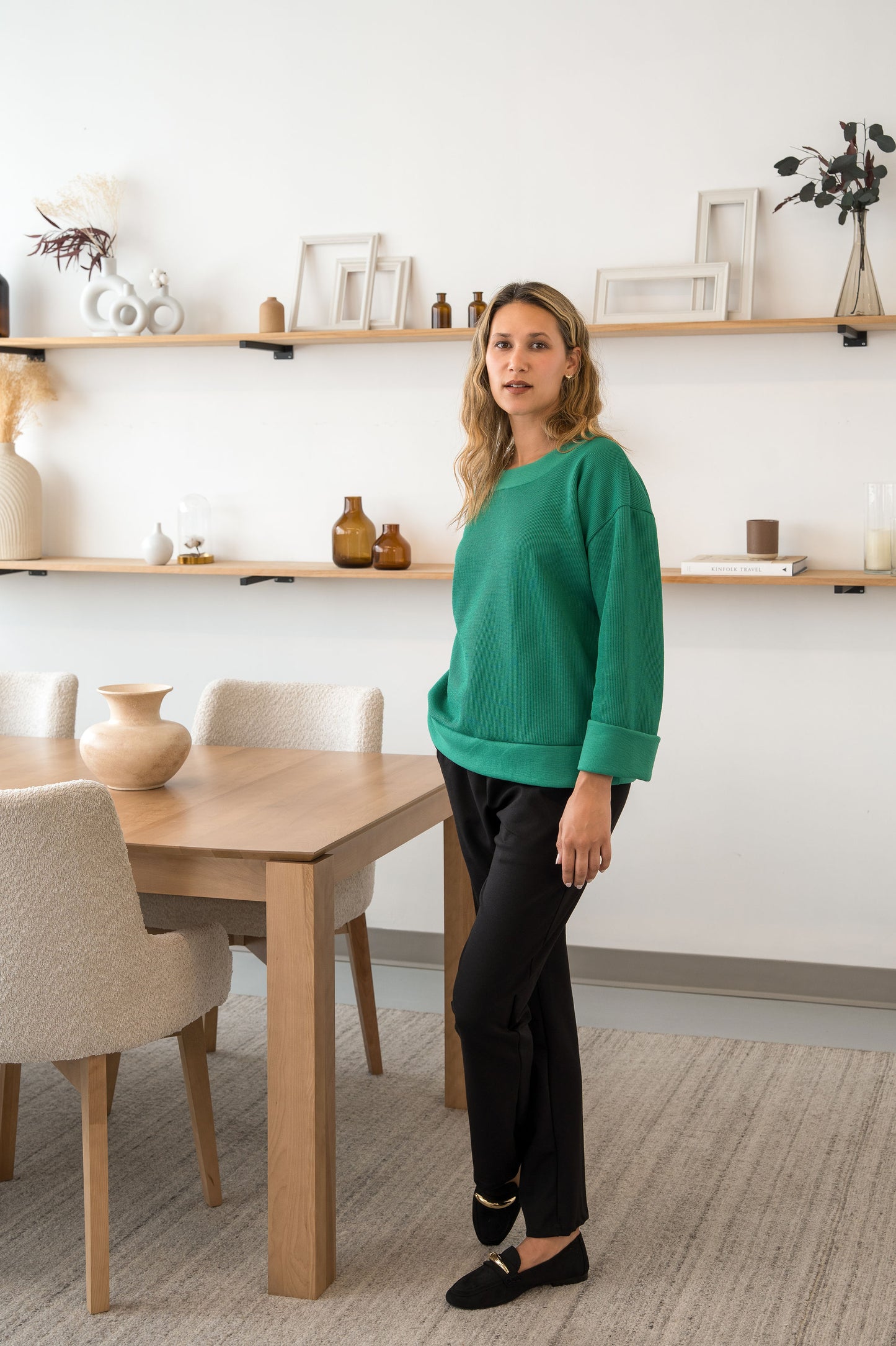 A woman wearing the Manhattan Pants by Cherry Bobin in Grey with the Mocha Sweater in Green and standing in a room with shelves.