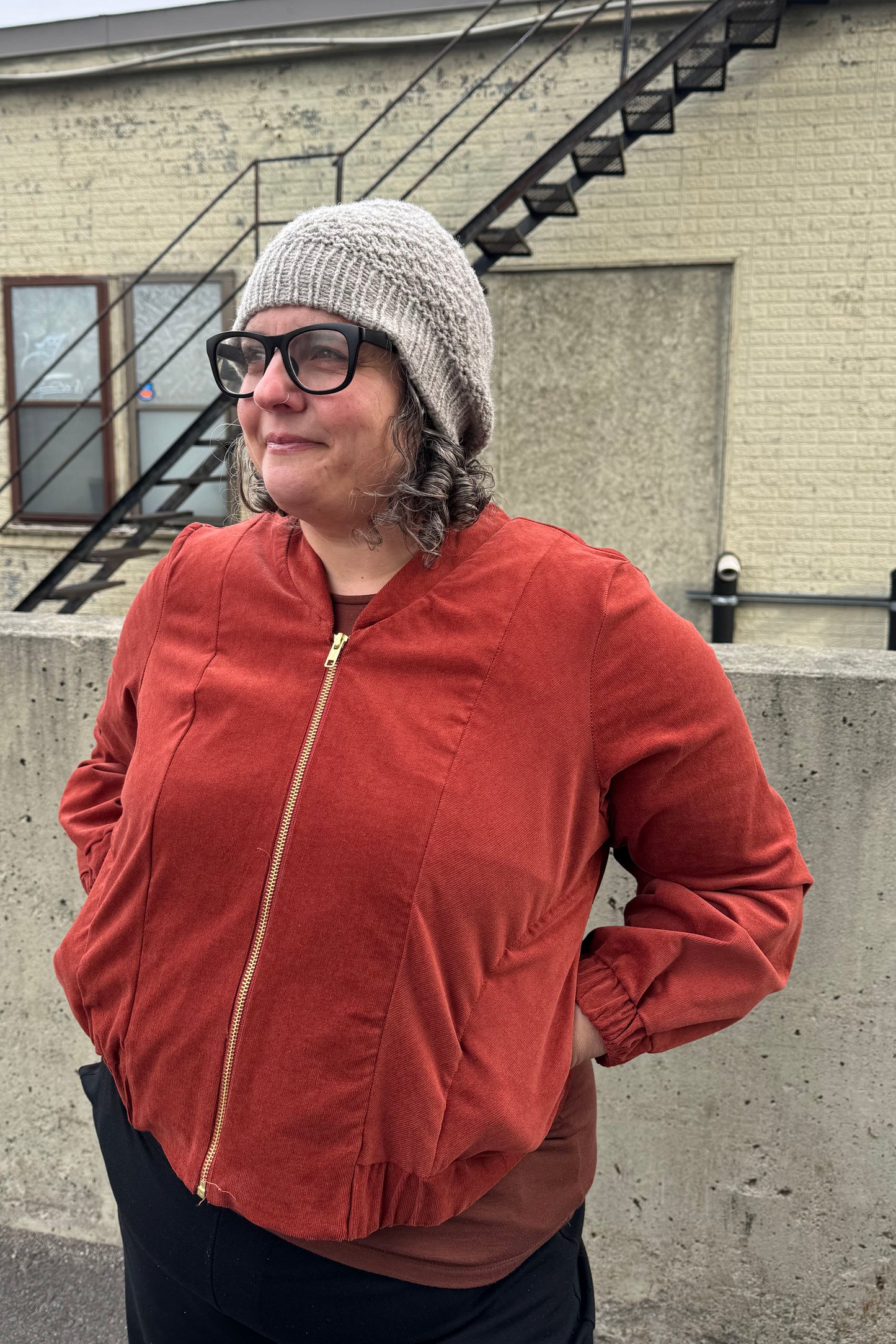 A woman wearing the Beret by Charlevoix Pure Laine in Grey with the Redrawn Zip Jacket by Eve Lavoie in Terracotta Corduroy. She is standing outside.